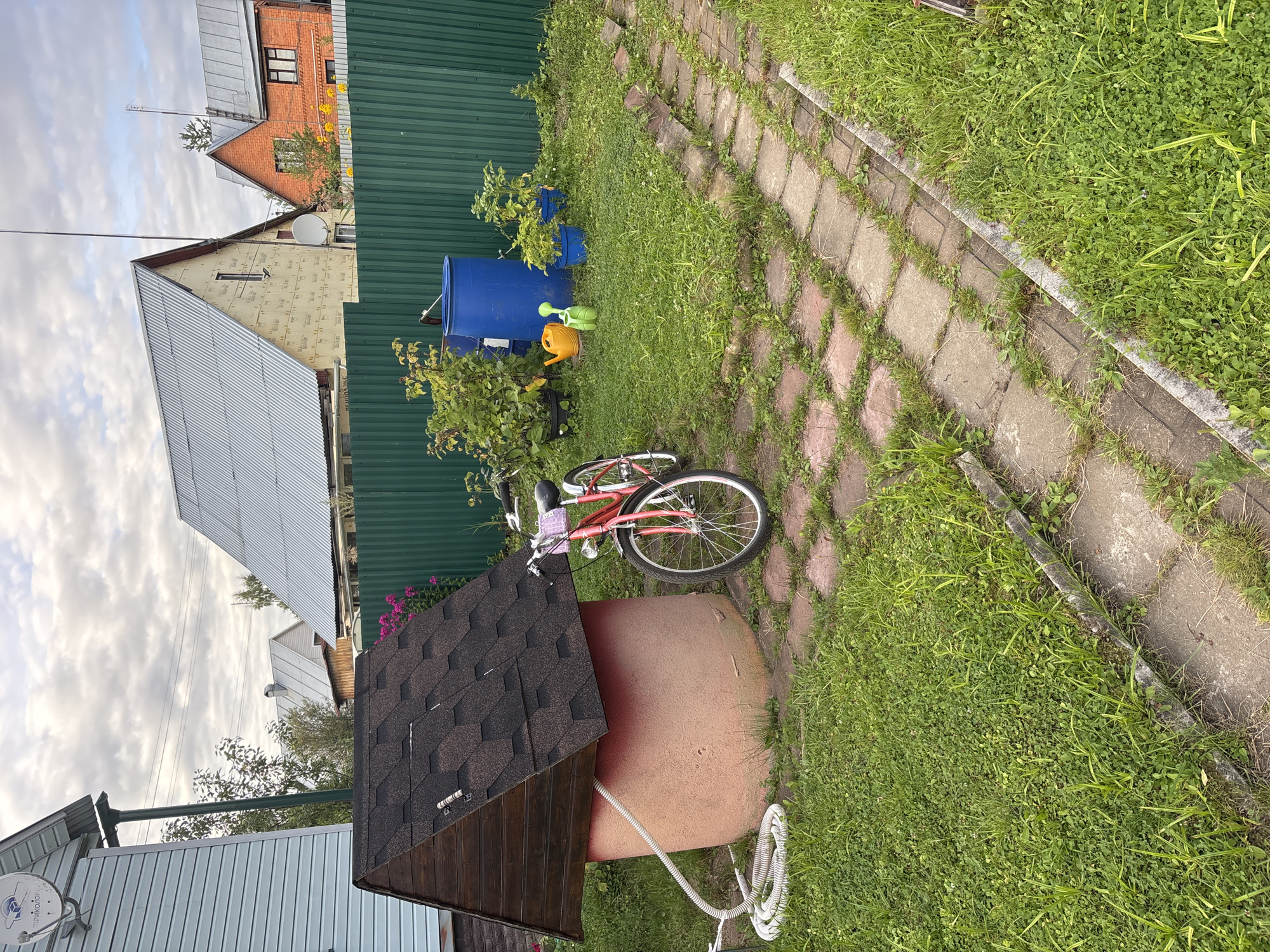 In the village of Nizinka SNT, Russia, a residential yard features a well with a dark shingled roof situated on a pinkish-orange concrete base. A white coiled hose extends from the well. To the right of the well, a pink bicycle with a wicker basket is parked. Beyond the well and bicycle, a green corrugated metal fence encloses the yard. In front of the fence, a large blue plastic barrel stands beside a cluster of green bushes and potted plants. Two watering cans, one yellow and one green, are placed on the grass. A paved path made of square reddish-brown pavers runs from the foreground towards the right of the image. The yard is covered in green grass and weeds. In the background, several houses with metal roofs and a satellite dish are visible under a cloudy sky.