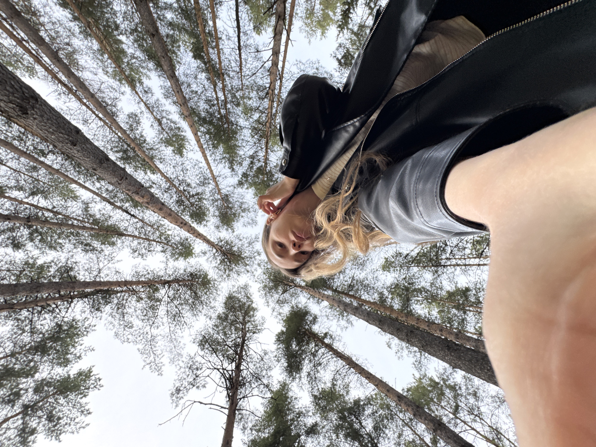 A low-angle shot of a woman standing in a forest, potentially in Прикамский, Russia. She is wearing a black leather jacket and is looking down at the camera. The trees in the forest are tall and dense, with bare branches. The sky is overcast and grey. The woman's hand is near her face, possibly adjusting her hair.