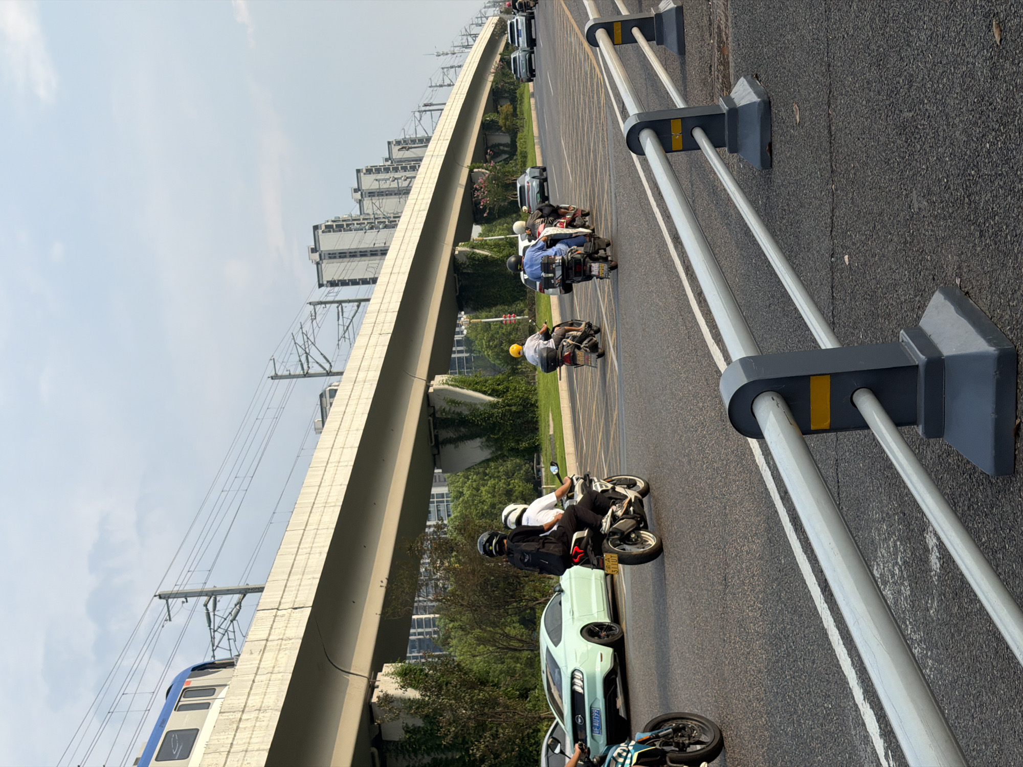 An elevated railway track runs horizontally across the frame, supported by concrete pillars, above a multi-lane road. A train is partially visible on the track. Several vehicles are on the road, including a light green sports car, multiple motorcycles with riders, and other cars in the distance. The motorcycles are traveling in the same direction as the sports car. Buildings and trees are visible beyond the road. The scene appears to be in Jiangjin, China, based on the presence of an elevated railway and the general urban environment.