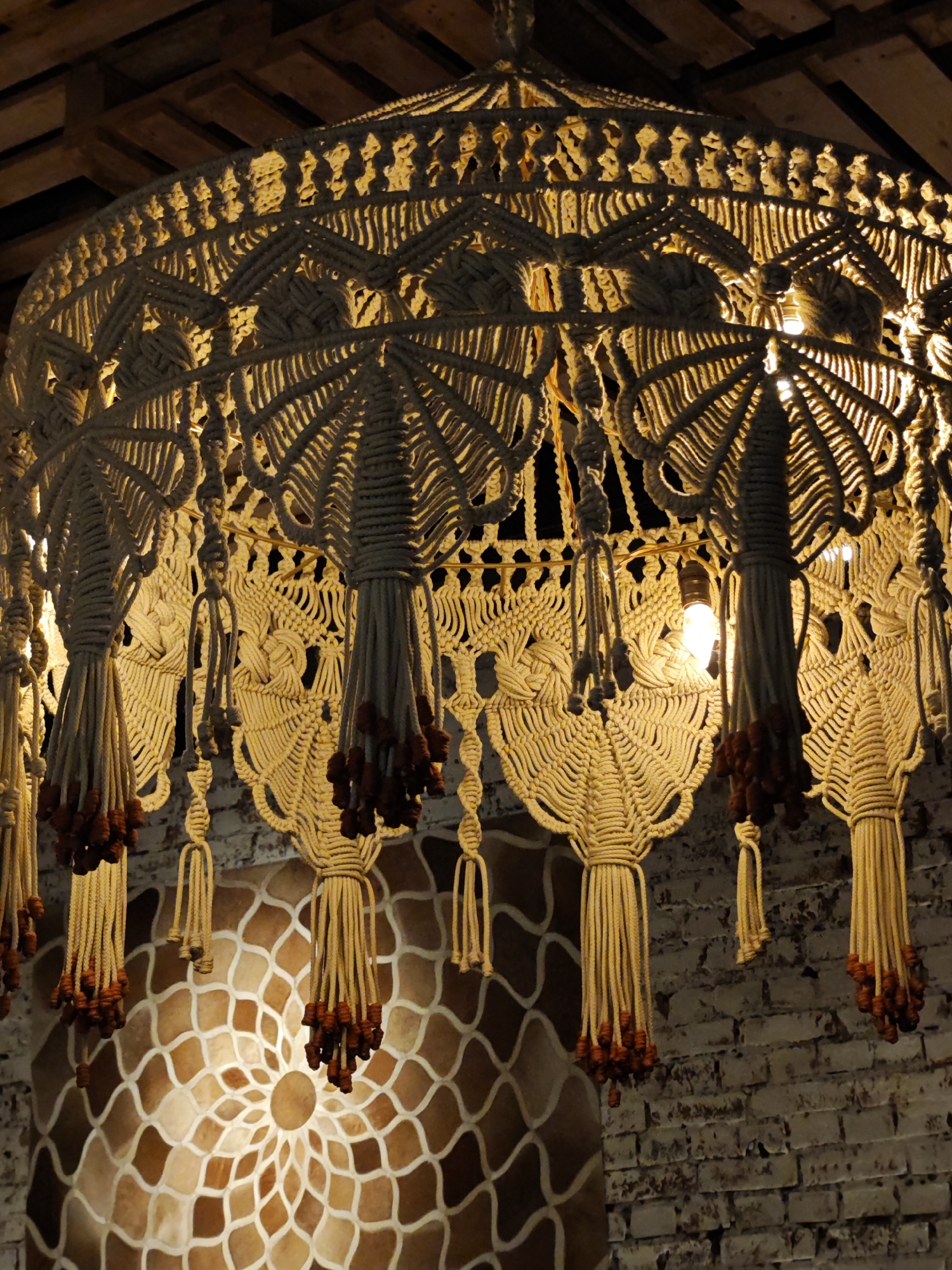 The image displays a macrame chandelier illuminated by incandescent bulbs, hanging from a wooden ceiling. The chandelier is intricately woven and features decorative tassels with dark bead accents. Below the chandelier, a patterned wall with a circular motif is visible, and to the right, a brick wall serves as the backdrop. The overall scene suggests an interior setting, possibly a cafe or restaurant in Saint Petersburg, Russia, characterized by its bohemian or rustic decor. There are no people or animals present in the image.