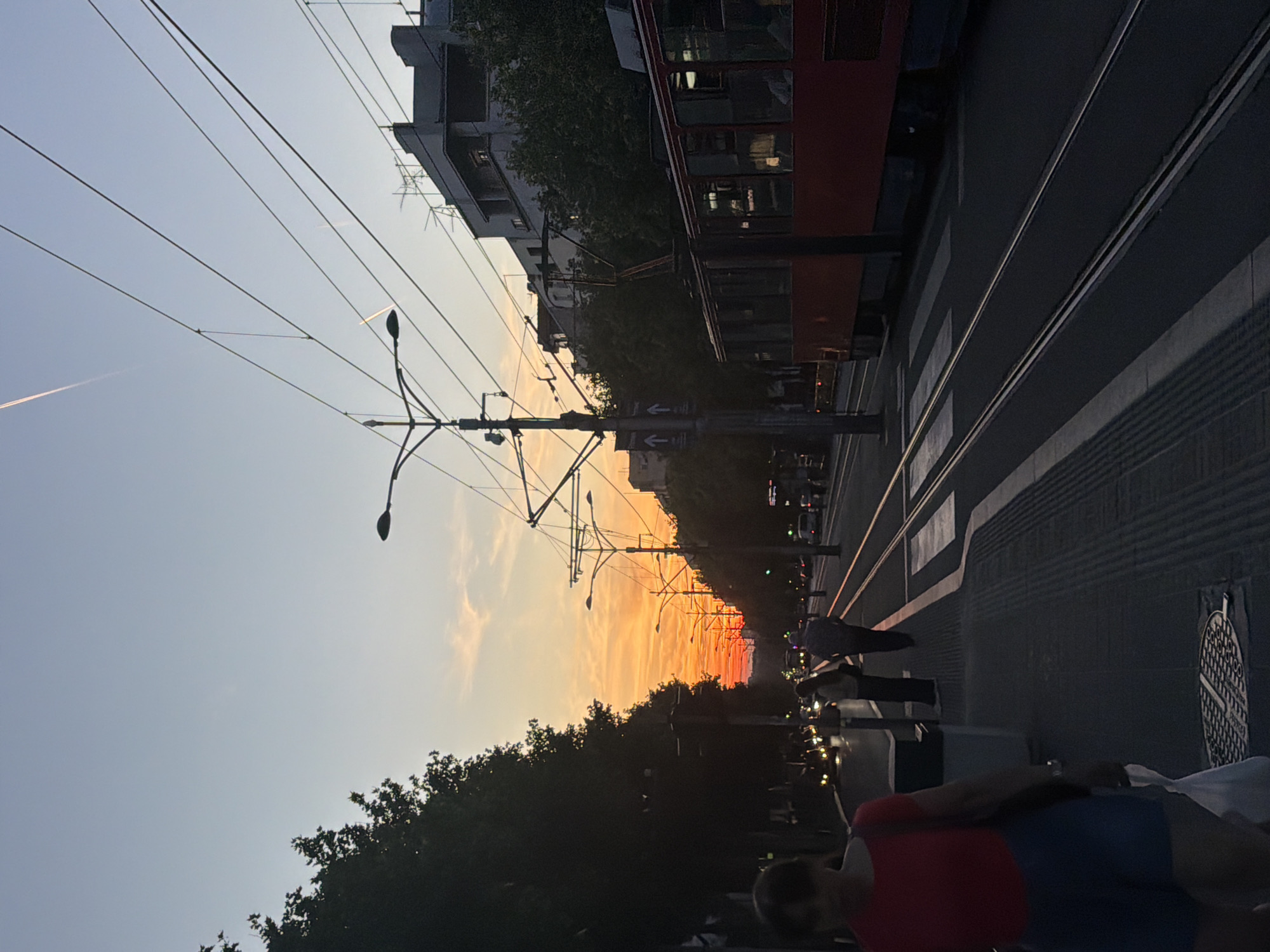 This image captures a vibrant urban street scene at sunset in Zvezdara, Serbia. The sky dominates the upper half, transitioning from a clear light blue at the top to striking orange and red hues near the horizon, indicative of the golden hour. A faint airplane contrail is visible in the upper left portion of the sky.
In the midground, a complex network of overhead power lines and support poles for a tram system crisscrosses the sky. Multiple streetlights are attached to these poles. On the left side of the street, multi-story buildings are partially obscured by a dense line of trees, which appear as dark silhouettes against the bright sky. A red tram is visible on the tracks alongside these buildings, its windows reflecting the warm light.
The street features two sets of parallel tram tracks extending into the distance. Pedestrian crossing stripes (zebra crossing) are visible on the road surface. On the right side of the street, several people are present. In the immediate foreground, a person wearing a red shirt and blue shorts is seen from behind, partially silhouetted. Further back, other figures walk along the sidewalk or stand near the road, also appearing as silhouettes. One person carries a white bag. A circular manhole cover with an intricate pattern is visible on the paved surface. Blurry vehicle lights in the distance suggest ongoing traffic.
On a pole in the middle of the street, a small sign with two horizontal arrows pointing in opposite directions is visible. The image conveys the tranquil beauty of an evening in a bustling city environment.