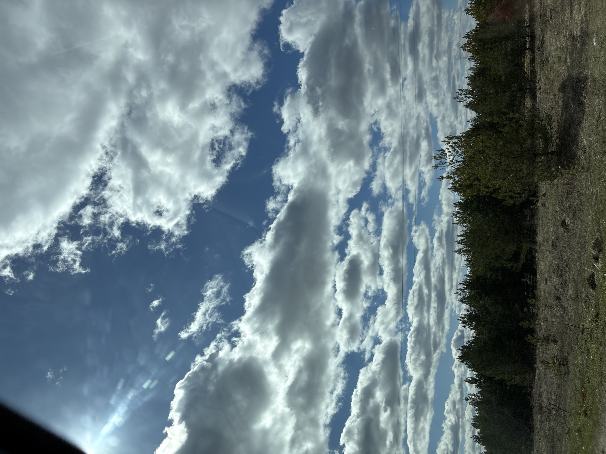 The image shows a view of a blue sky with scattered clouds, partially obscured by trees and terrain on the right side. The scene seems to have been captured from a moving vehicle.
The location is unknown, and there is no information about people, subjects, objects, actions, or interactions, only the landscape and sky are visible in the image.
