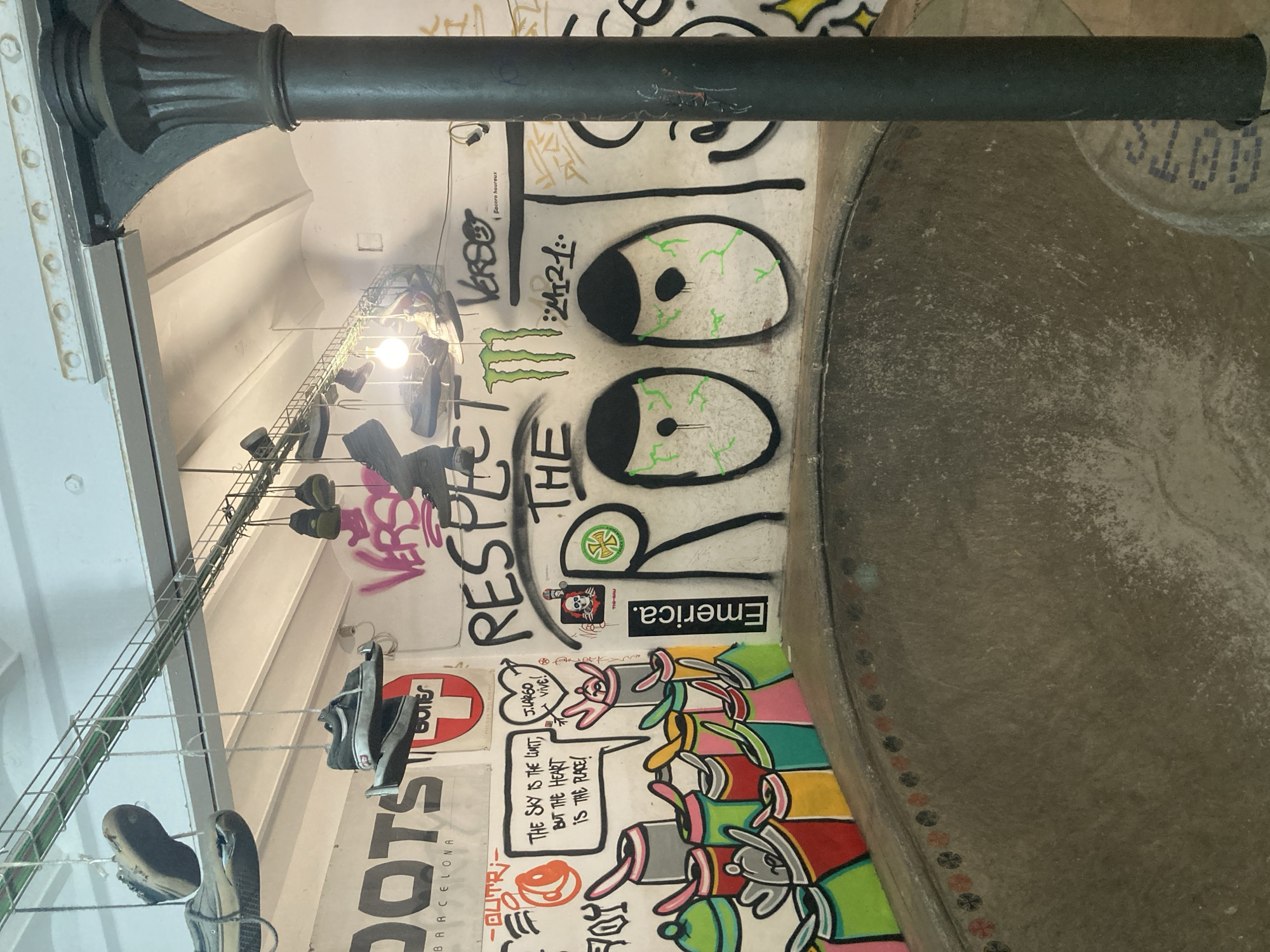 This image captures an indoor urban environment, likely a skatepark or a store with a skate culture theme, located in Barcelona, Spain. A prominent, dark, fluted metal column stands on the left, supporting the structure above. The walls are extensively covered in graffiti and street art, showcasing a vibrant and edgy aesthetic.
On the main wall, large cartoonish eyes with green irises and veins are painted, next to a "Monster" energy drink logo. Below this, the words "RESPECT THE ROOTS" are partially visible in stylized lettering. Further down, a colorful mural depicts a row of cartoon rabbits, some of which are conversing in speech bubbles. One bubble reads, "THE SKY IS THE LIMIT, BUT THE HEART IS THE PLACE!", and another nearby says "J. LARGO Vive!". Other visible text includes "Emerica." (a skate brand), "DOTS BARCELONA" on a poster, "VERSO", "Encore heureux", and various other tags and logos, including a red Swiss cross-like emblem and a "BONE$" logo.
To the right, a smooth, grey concrete surface curves upwards, forming part of a skate bowl or ramp. Its edge features a decorative border of small, patterned circular tiles. Hanging from the ceiling on the left, a metal grid display holds several pairs of shoes, illuminated by bare light bulbs, suggesting a retail or display purpose. The overall atmosphere is energetic and counter-cultural, typical of skate or streetwear venues. The indoor setting makes the time of day and weather indeterminate.