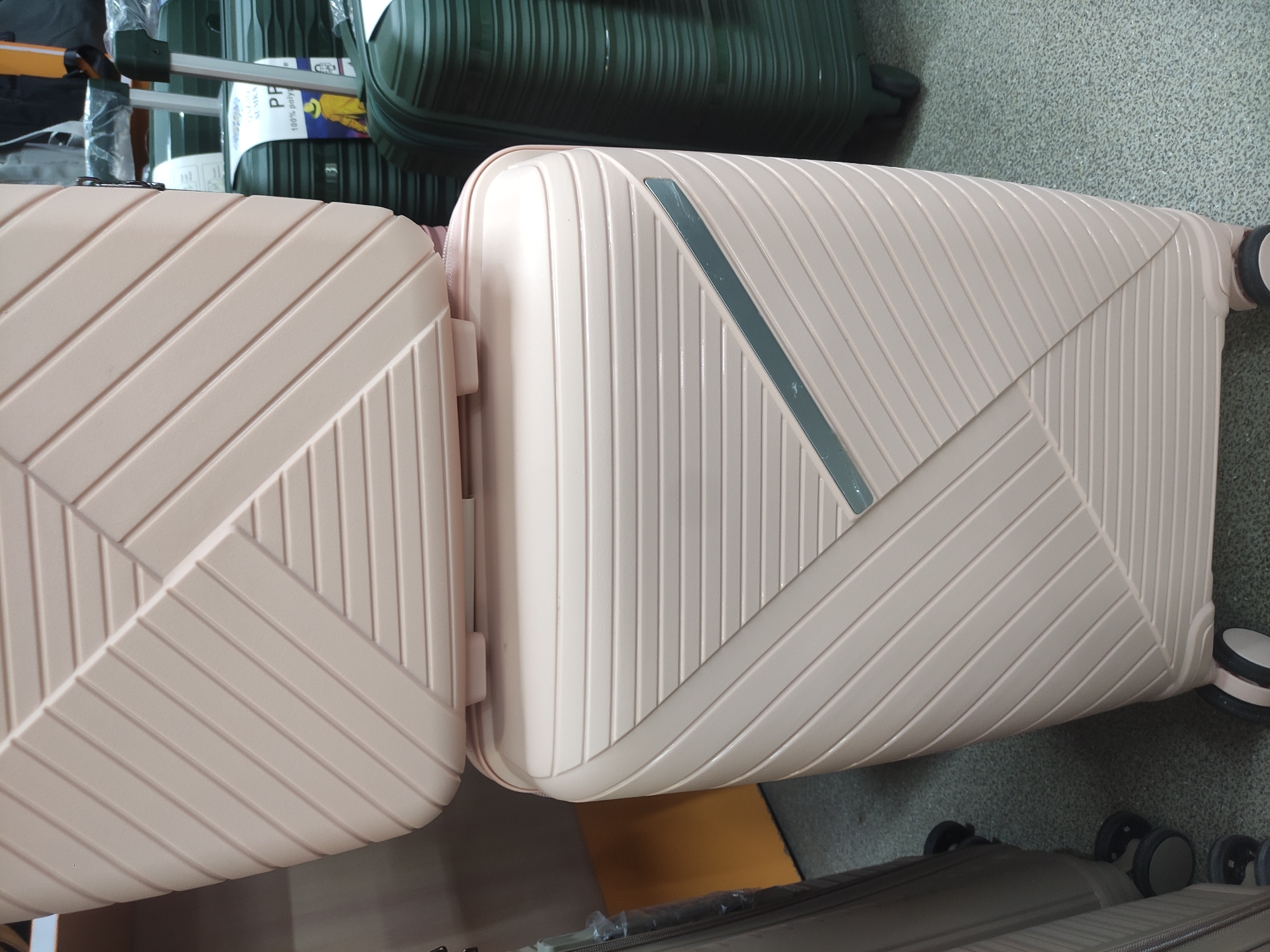 The image shows a close-up of stacked luggage, primarily a light pink suitcase on top of another similar suitcase. The luggage has a ribbed texture and a geometric pattern. To the right of the pink luggage, a dark green suitcase is visible. The floor appears to be carpeted. The scene is indoors, likely in a retail setting. There is no visible indication of the city of Oral, Kazakhstan within the image itself or the provided crops.