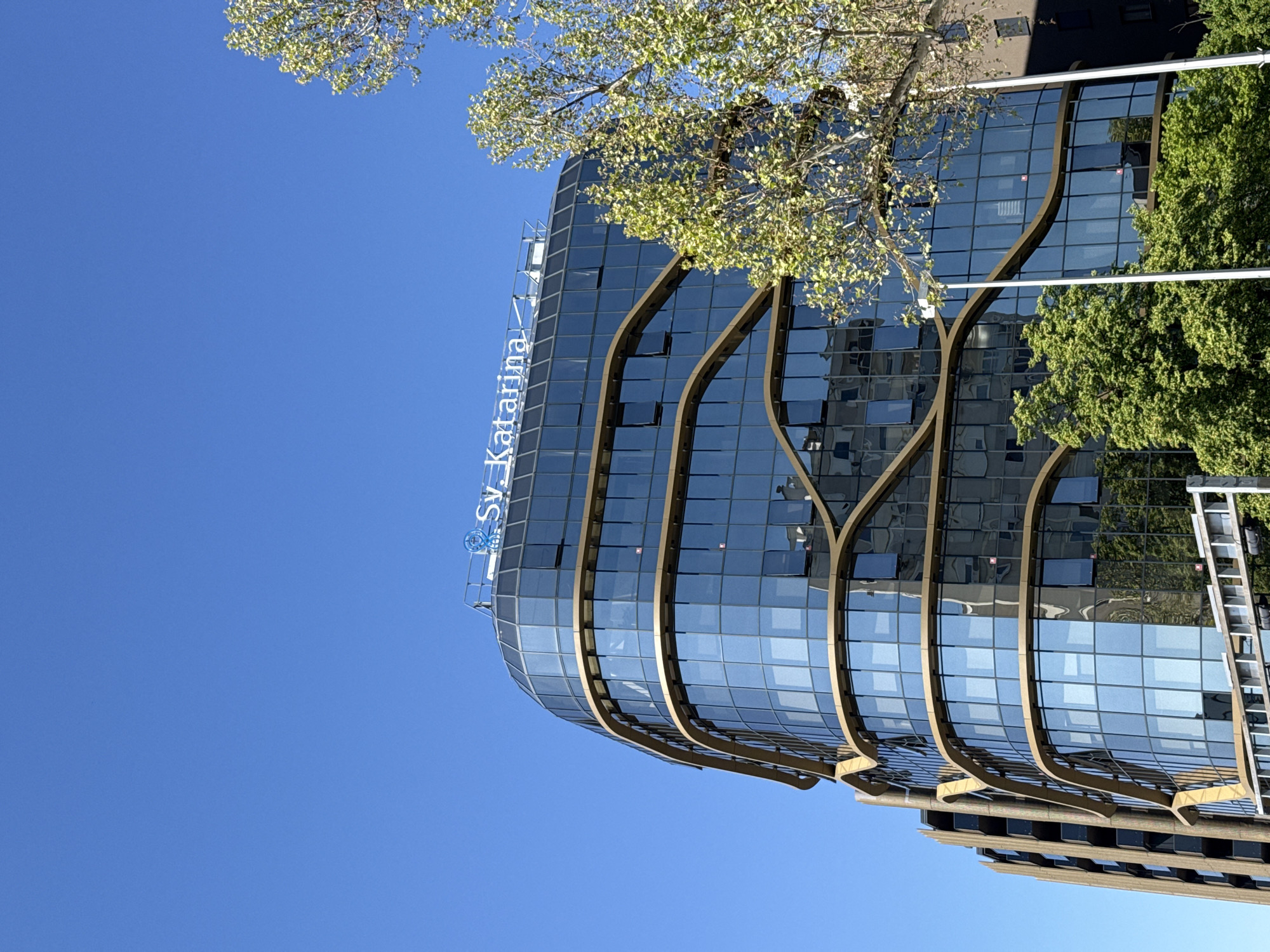 The image shows the Sv. Katarina Specialty Hospital in Zagreb, Croatia. The building is a modern glass structure with curving, decorative exterior elements. Part of the building's name is visible on the facade. The sky is clear and blue. A tree with light green leaves is partially visible, obscuring parts of the building. Another green tree is visible to the right. Parts of surrounding buildings are also visible.