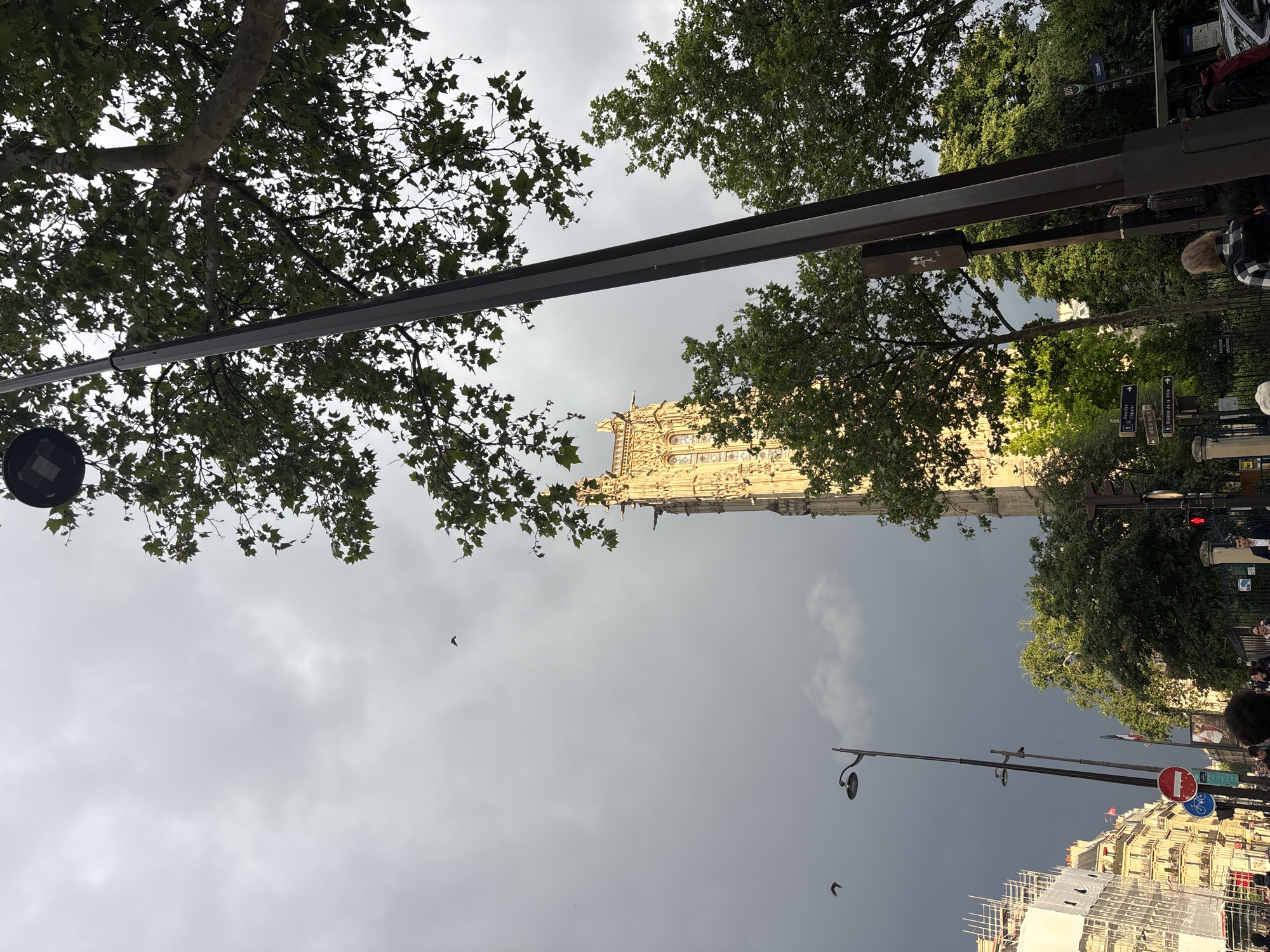 The image shows the Saint-Jacques Tower in Paris, France, as seen from below on a cloudy day. The tower is framed by trees and a metal structure. People are present on the right side of the image, with buildings visible in the background. Street signs and lampposts are also visible. Birds are seen flying in the sky. Scaffolding is present on the building below.