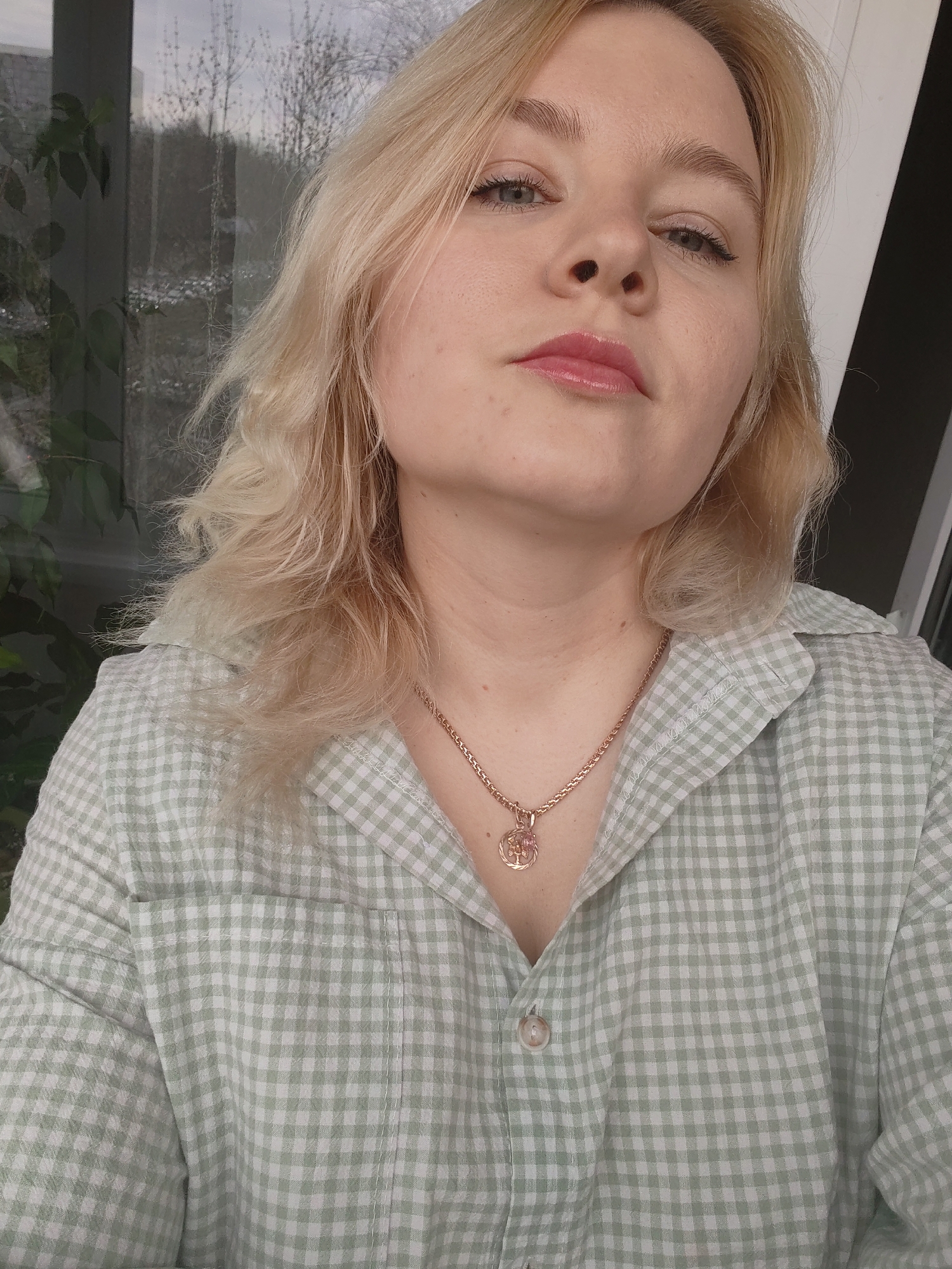 A medium shot depicts a blonde woman with fair skin, gazing downward toward the camera. She wears a green and white checkered shirt. A thin gold chain necklace with a pendant sits at the base of her neck. The background reveals a glimpse of a window with greenery visible outside. The location is Izhevsk, Russia.