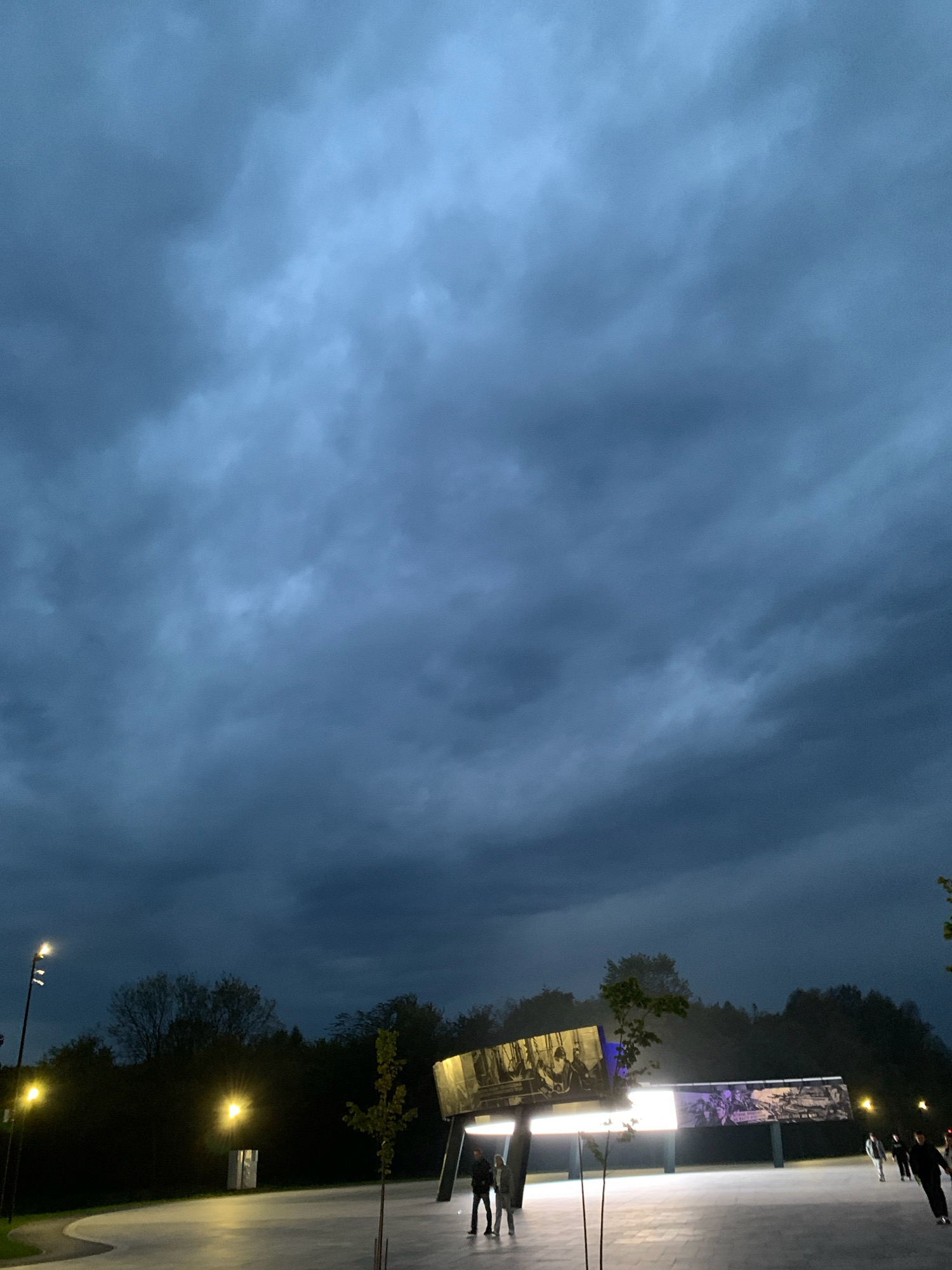 The image depicts an outdoor scene at dusk, characterized by a dark, cloudy sky overhead. In the mid-ground, illuminated structures with large display panels are visible, showcasing images or text. Several people are present, some walking and others standing. The foreground shows a paved area with lighting poles and young trees. The overall atmosphere is dimly lit, suggesting the transition from daylight to night. The location is identified as Prokopyevsk, Russia.