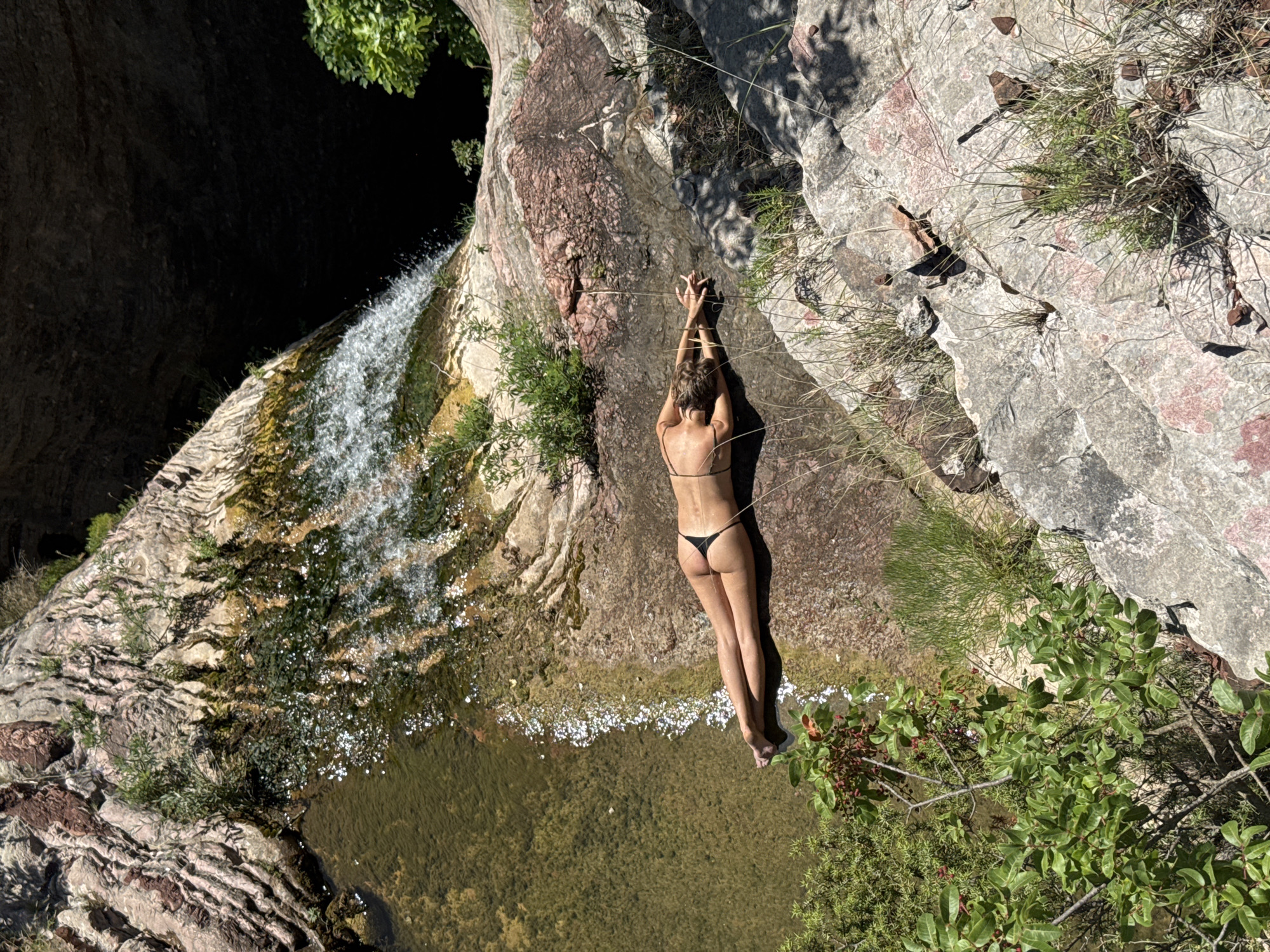 A person in a black bikini lies on a rocky ledge next to a waterfall. Their arms are raised and clasped overhead, forming a heart shape with their fingers. The person's back is to the camera, and they are facing a pool of water below the waterfall. The scene is bathed in sunlight, suggesting it is daytime. The surrounding rocks are rough and textured, with patches of green vegetation growing in crevices. The water of the waterfall appears clear and refreshing, cascading down the rocks into the pool. The setting appears to be a natural, outdoor environment, possibly a secluded spot in a canyon or gorge. The overall mood is one of relaxation and enjoyment of nature. There is no visible text in the image.