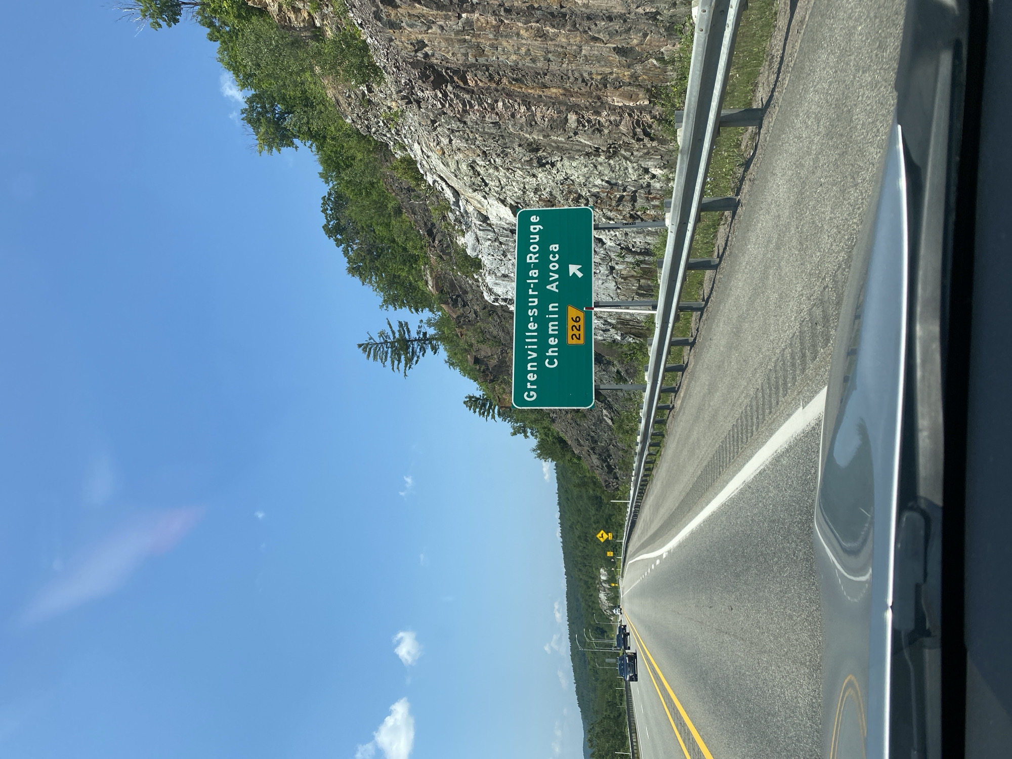 The image depicts a highway scene on a sunny day. A green highway sign indicates "Grenville-sur-la-Rouge Chemin Avoca" with a route number "226" and an arrow pointing to the right. The sign is positioned on the right side of a multi-lane highway. The highway runs through a landscape with rocky cliffs and lush green trees on the right, and a forested hillside on the left. Several cars are visible on the road. The foreground shows the hood and windshield of a vehicle, suggesting the photo was taken from a car. The sky is blue with some scattered clouds. The location is identified as Pointe-au-Chêne, Canada.