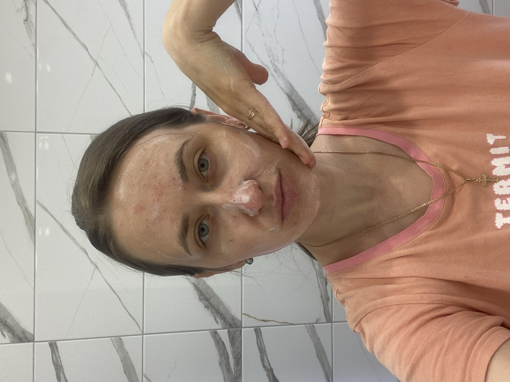A woman is taking a selfie in a bathroom with tiled walls. She has light-colored skin and blue eyes, with her hair pulled back. She is wearing a peach-colored t-shirt with a pink trim and a gold necklace with a cross pendant. She has a facial mask on her face, with foamy white cream around her nose and mouth. Her right hand is touching her face, with a ring on her index finger. The woman appears to be in her late 20s or early 30s.