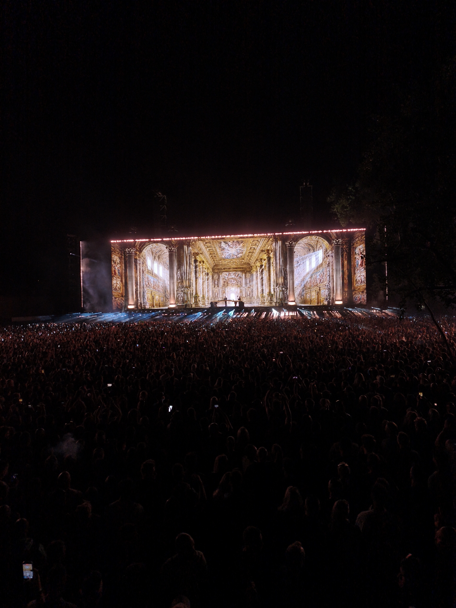 An outdoor concert is taking place at night. A massive crowd of people is gathered in front of a large stage. The stage features an elaborate, illuminated backdrop that resembles a grand interior with columns, arches, and detailed artwork, possibly a projection. Bright stage lights are directed towards the audience, creating beams of light that cut through the dark. Smoke or fog is visible rising from the stage. Many audience members are holding up their phones, likely recording or taking pictures of the performance. The scene is dark, with the primary light sources being the stage and the screens. The overall atmosphere is one of excitement and engagement, typical of a live music event.