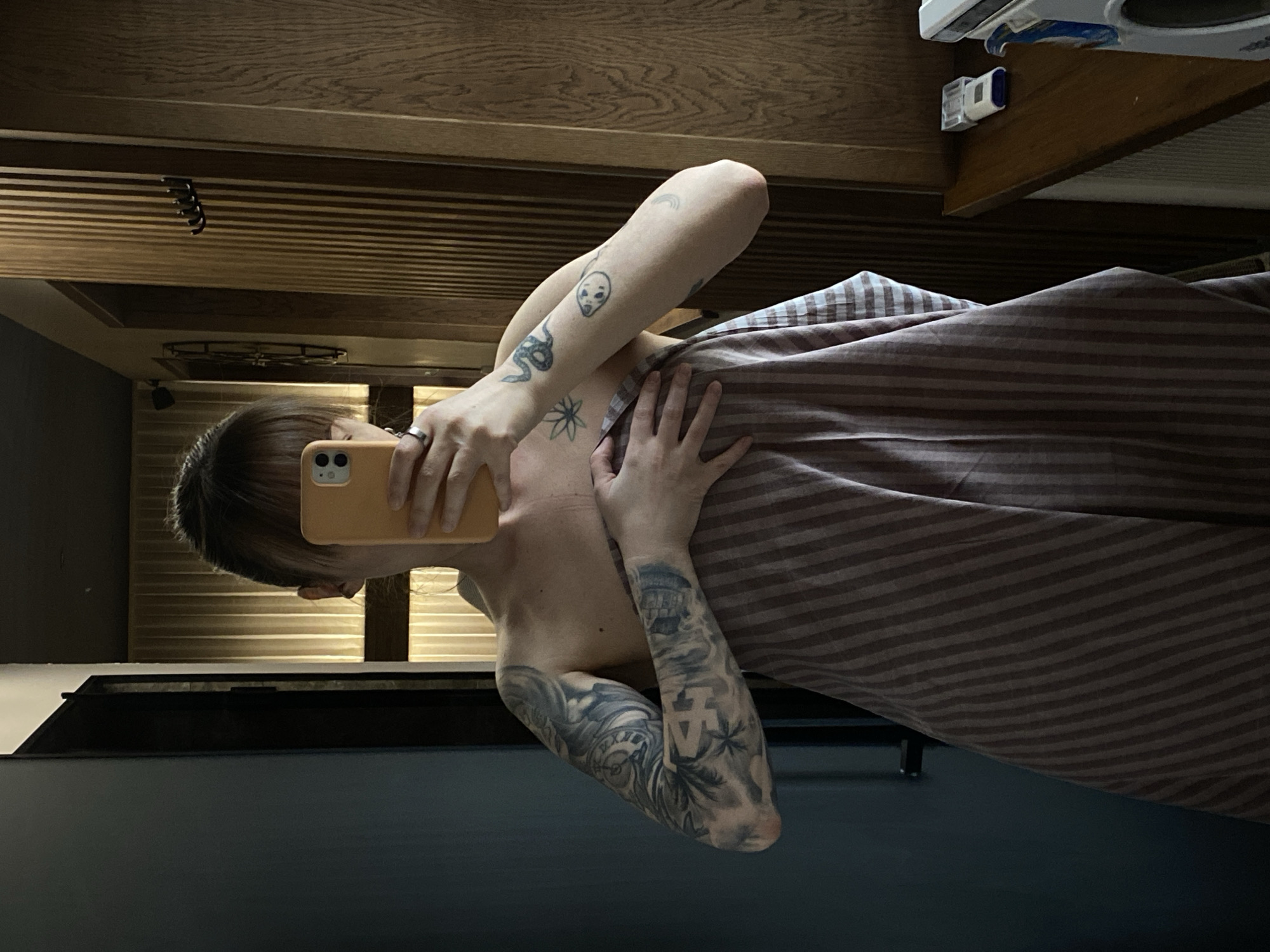 A person with tattoos is taking a selfie in a room. The person is partially covered by a striped blanket. The background includes dark walls, wood-paneled ceiling elements, and miscellaneous objects on a shelf. It is impossible to determine if the picture was taken in Votkinsk, Russia.