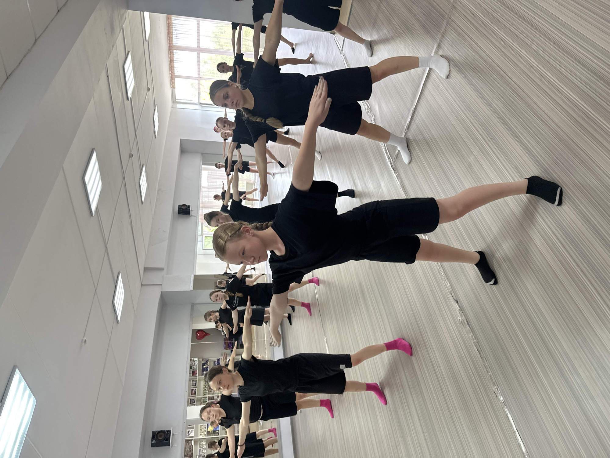 In a dance studio in Votkinsk, Russia, a group of young girls, likely students, are engaged in a synchronized movement exercise. They are all dressed in black t-shirts and shorts, with some wearing brightly colored socks, such as pink or white. The room features a polished wooden floor, large mirrors lining the walls, and bright fluorescent lighting panels on the ceiling. The girls stand in rows, their arms extended horizontally to their sides, with their bodies positioned in a stable stance. The focus of the image is on the central figures, particularly the girl in the foreground on the right, whose gaze is directed forward with a serious expression. In the background, other girls are visible performing the same exercise, suggesting a class or rehearsal setting. The mirrors reflect the entire scene, doubling the visual of the participants and the studio's spaciousness.