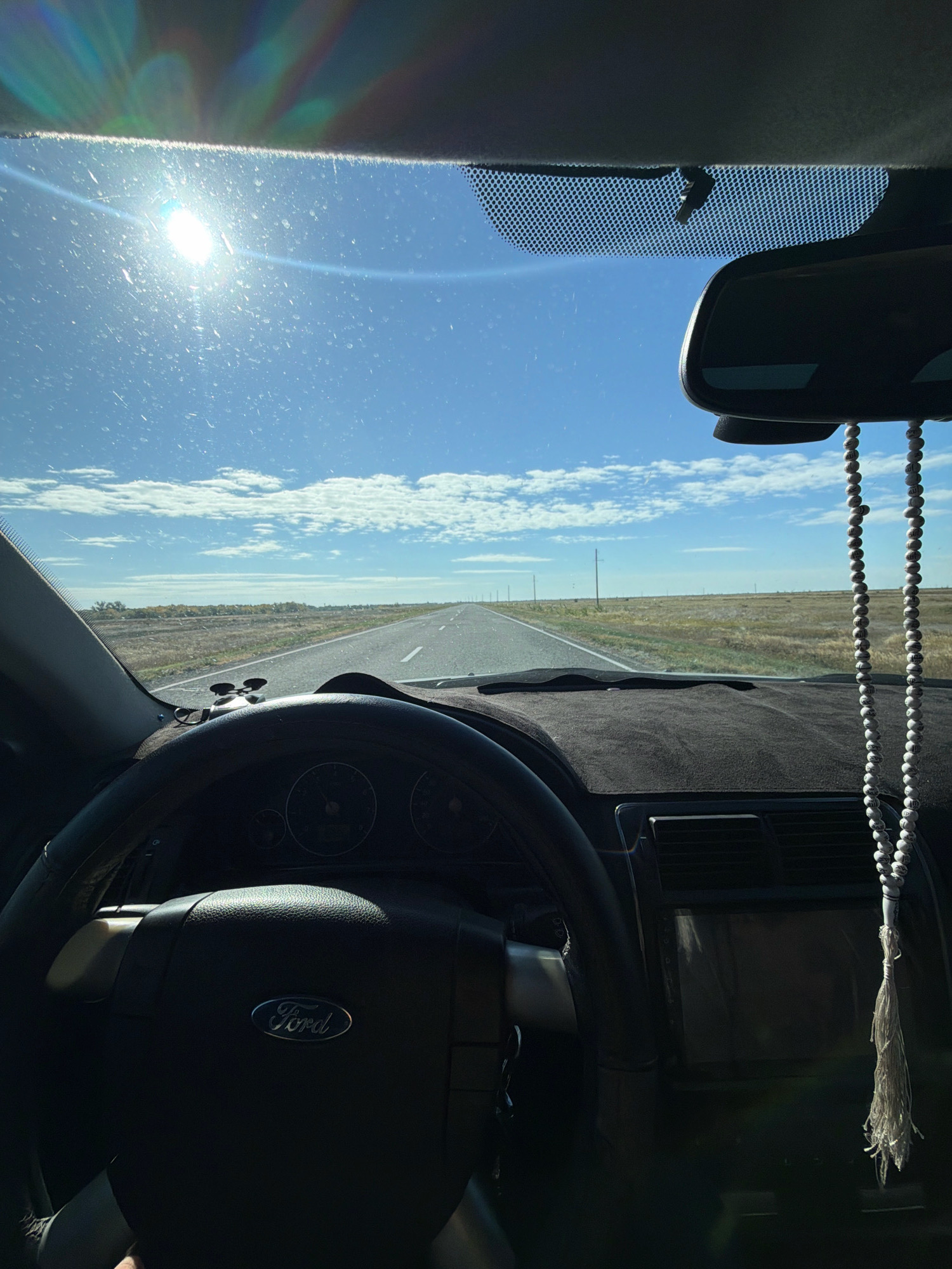 The image is a first-person perspective from inside a car, looking out through the windshield at a straight, paved road stretching into the distance under a bright blue sky with scattered clouds. The sun is visible, creating lens flare and glares on the windshield. The interior of the car shows a black dashboard, a steering wheel with a Ford logo, and a rearview mirror with prayer beads hanging from it. The landscape outside the car is flat and rural, with dry grass and some distant trees or structures. The road appears to be in a sparsely populated area, possibly in the city of Көктүбек ауылдық округі, Kazakhstan, given the vast, open terrain.