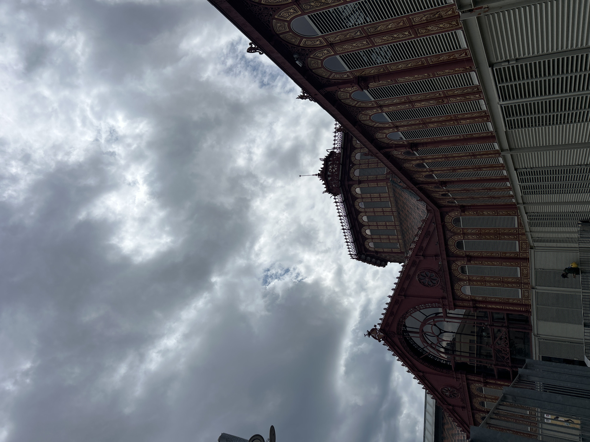 The image shows an upward angle view of a building facade against a cloudy sky. The building has a red and gold decorative exterior with arched windows and intricate patterns. The design suggests it might be a historic market or public building. Based on architectural style and location clues, it is likely the Mercat del Born (Born Market) in Barcelona, Spain. The market features a combination of ornate metalwork and functional architectural elements like the grey ventilation system.