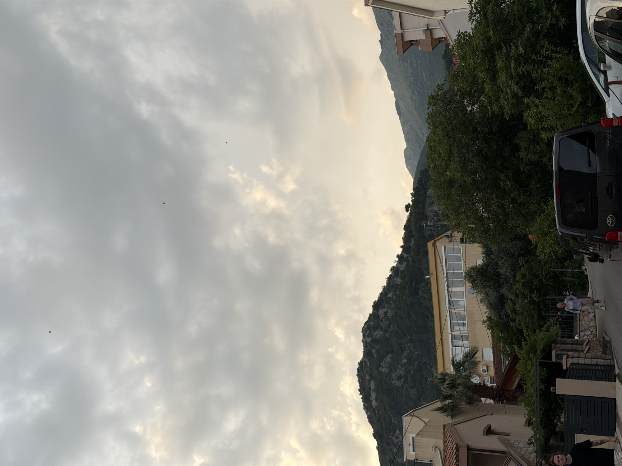 The image depicts a street scene in Budva, Montenegro, with a view of residential buildings and a forested mountain range in the background under a cloudy sky. In the foreground, a dark-colored van is parked on the right side of the road. A white car is parked next to it, partially visible. On the left side of the street, a man wearing a black t-shirt and white shorts is walking away from the camera. A woman wearing a light-colored shirt and light-colored pants is walking towards the camera on the right side of the road. A few small, dark objects, possibly birds, are visible in the sky. The architecture includes buildings with terracotta roofs and yellow facades, interspersed with dense green foliage and a palm tree. The overall lighting suggests it is late afternoon or early evening.