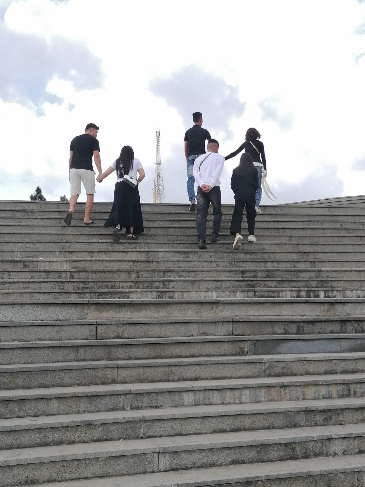 The image shows a group of six people ascending a wide, concrete staircase outdoors. The sky above is overcast with scattered clouds. The people, viewed from behind, are walking up the steps in varying formations. In the foreground, a man in a black t-shirt and white shorts walks hand-in-hand with a woman in a white t-shirt and long black skirt. Behind them, a man in a black t-shirt and jeans walks, followed by a man in a white long-sleeved shirt and dark jeans. Further up the stairs, a woman in a black jacket and blue jeans is trailed by another woman in black pants and a black top. A tall, slender tower, possibly a communication tower or a landmark, is visible in the background, partially obscured by trees on the left. The staircase appears to be part of a public space, possibly a monument or a viewpoint in Da Lat, Vietnam, given the cloudy sky and the architectural style.