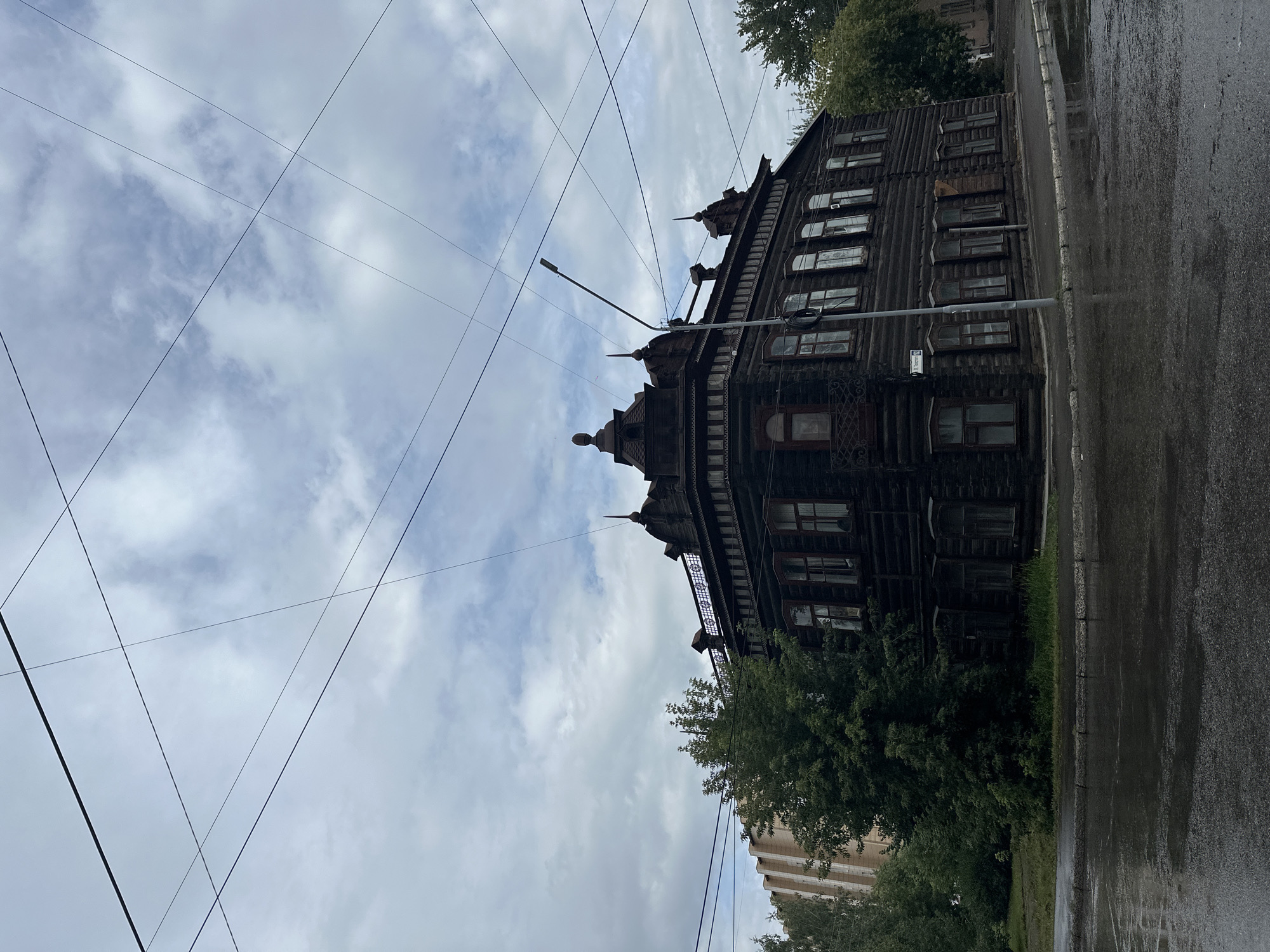 An aged, dark wooden two-story building with ornate architectural details stands on a street corner in Biysk, Russia. The building features multiple windows on both floors, some with decorative shutters. The roof is complex, with gables and finials. In front of the building, a streetlamp is visible. Overhead, a network of utility wires crisscrosses the cloudy sky. To the left of the wooden building, a modern, light-colored building is partially visible, and several green trees are present. The street and sidewalk in the foreground are wet, suggesting recent rain, with reflections visible on the asphalt. A street sign reading "ул. Цеховая 102" (Tsekhovaya Street 102) is affixed to a pole in front of the wooden building, indicating the specific location. No people are visible in the scene.