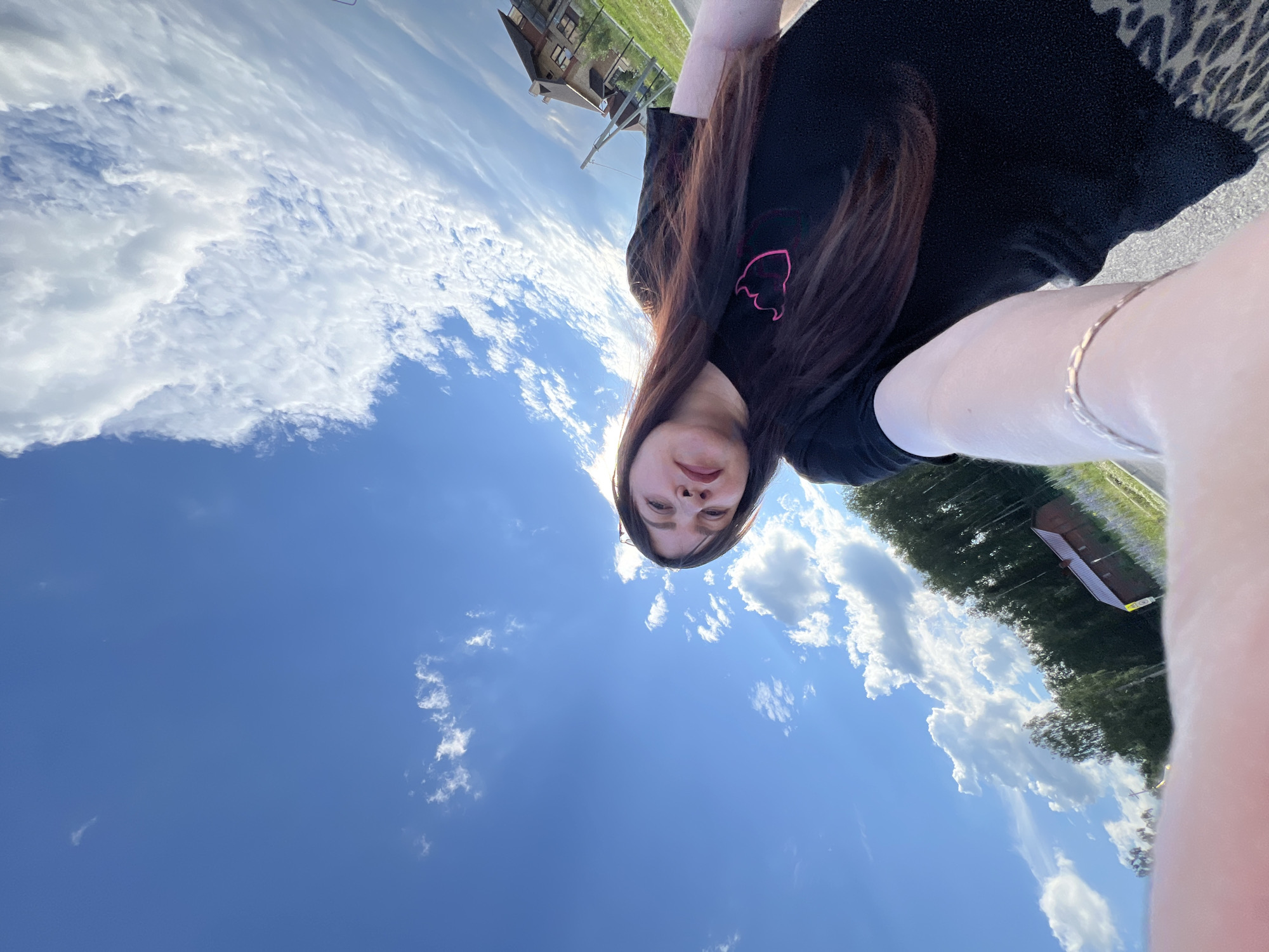 A selfie is taken outdoors on a sunny day with a bright blue sky filled with white, fluffy clouds. The subject, a woman with long, dark brown hair and fair skin, is looking up and slightly to the side towards the camera, with her mouth slightly open. She is wearing a black t-shirt with a pink outline of a heart on the left side of her chest, and a patterned skirt or pants with a leopard print design. A delicate silver bracelet is visible on her left wrist.
In the background, the scene appears to be a suburban or rural area. A modern, multi-story house with a gabled roof and several windows is visible on the right. To the left of the house, there is a dense line of tall, dark green trees. A small, reddish-brown wooden building with a dark roof is nestled amongst the trees. A thin, yellow sign is attached to a pole in front of the wooden building. The ground is covered with green grass, and a paved surface, possibly a road or sidewalk, is visible in the foreground. The location is identified as Noviy, Russia.