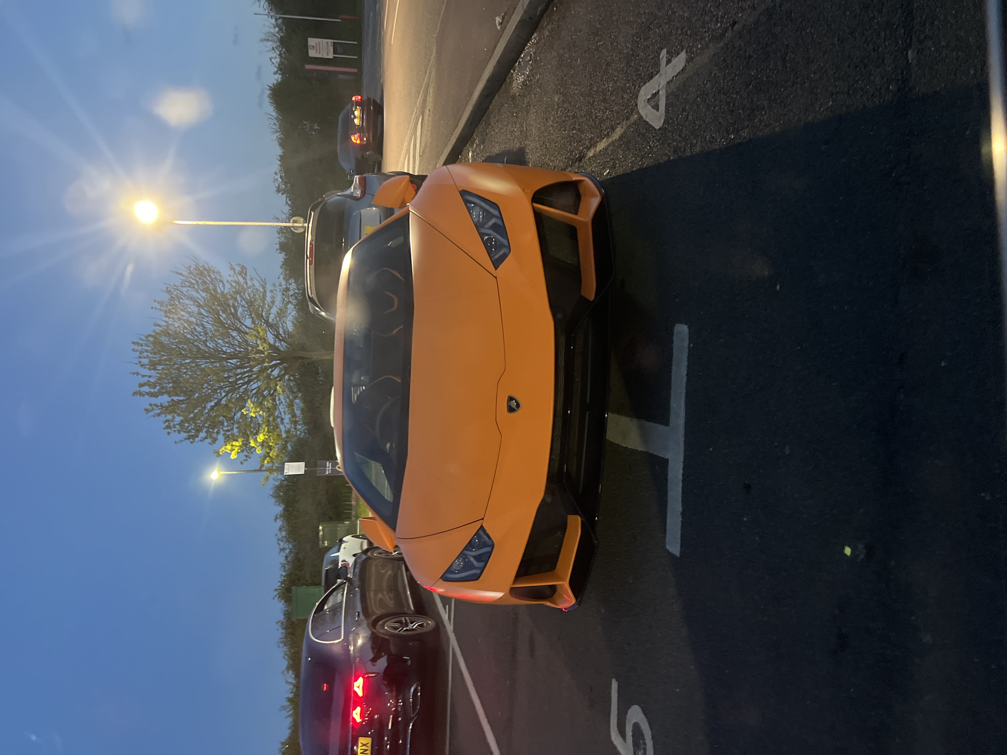 The image shows an orange Lamborghini parked in a parking space marked with an "8". A dark-colored sedan is parked behind it. A street lamp illuminates the area. The image was taken outdoors, at dusk or early evening. The location is in Gorleston-on-Sea, United Kingdom.