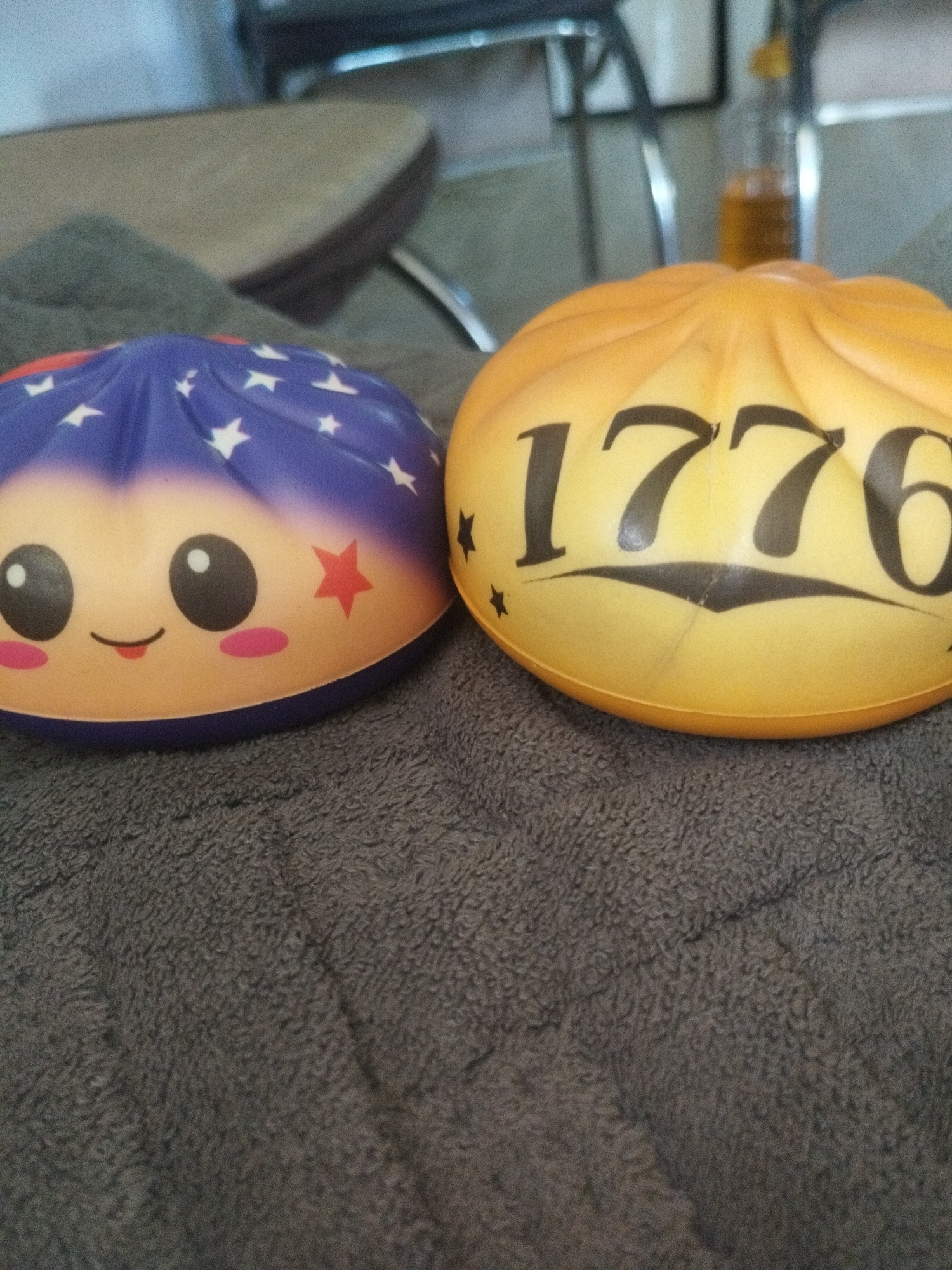 Two squishy toys are placed on a dark gray, textured surface. The toy on the left is blue with white stars and has a smiley face with two black eyes, pink cheeks, and a small red star on its right side. The toy on the right is yellow and has the numbers "1776" printed in black on its surface, along with some small black stars. The background is out of focus, showing hints of furniture and a bottle with liquid. There are no people visible in the image, and no context is provided to place it in Oskemen, Kazakhstan.