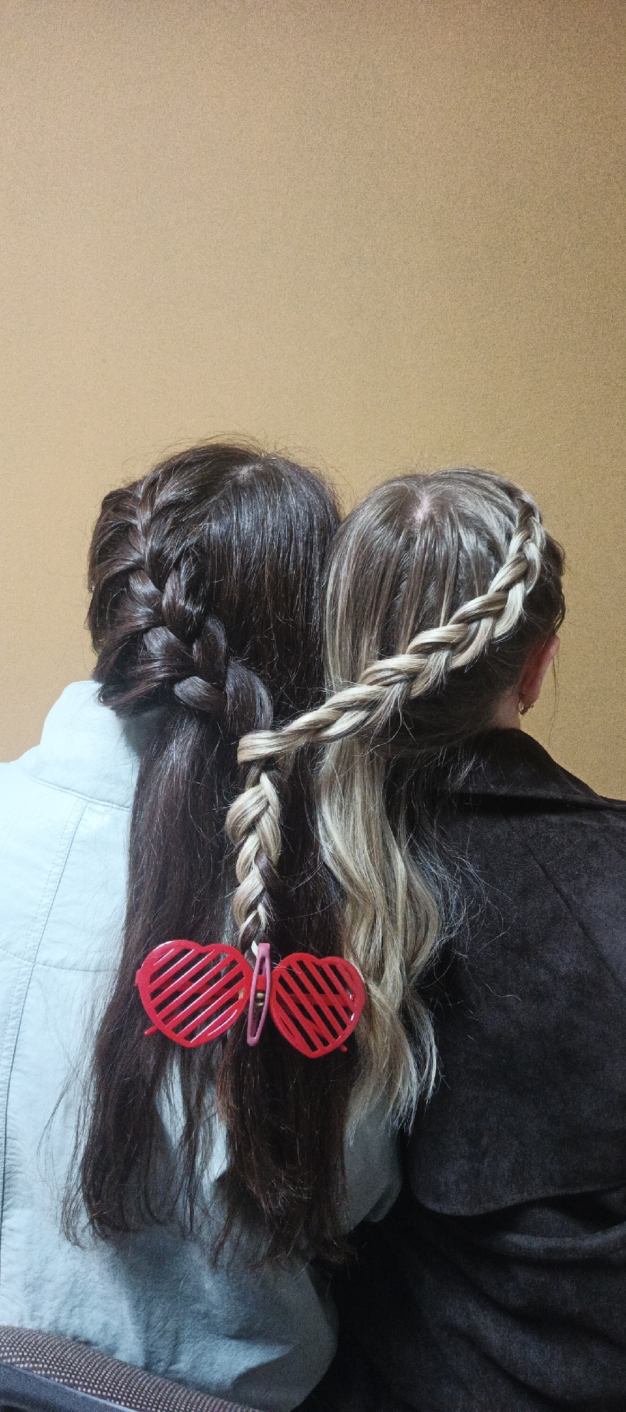 The image, taken in Chaikovsky, Russia, captures the backs of two individuals whose hair has been playfully styled to connect them. The subject on the left has dark brown hair, with a thick braid visible on the left side of their head, blending into a mass of long, straight hair. They are wearing a light blue jacket or shirt. The subject on the right has lighter, possibly highlighted, hair with shades of blonde and light brown. A single French braid starts from the top of their head and extends downwards. They are wearing a dark brown or black coat.
A unique detail is how their hairstyles intertwine: a portion of the lighter-haired individual's braid crosses over and is woven into a section of the darker-haired individual's loose hair. Both strands are then secured together near the bottom by a striking red, heart-shaped hair clip. This clip features a slatted design, resembling shutter shades. The overall arrangement suggests a close bond, friendship, or a fun, creative hairstyle project.
The setting appears to be indoors, against a plain, light brown or beige wall, providing a simple backdrop that highlights the intricate hairstyles and the connecting clip. There are no discernible location cues beyond the plain wall. No activity beyond posing for the picture is evident. The time of day cannot be determined from the indoor lighting. There is no visible text in the image.