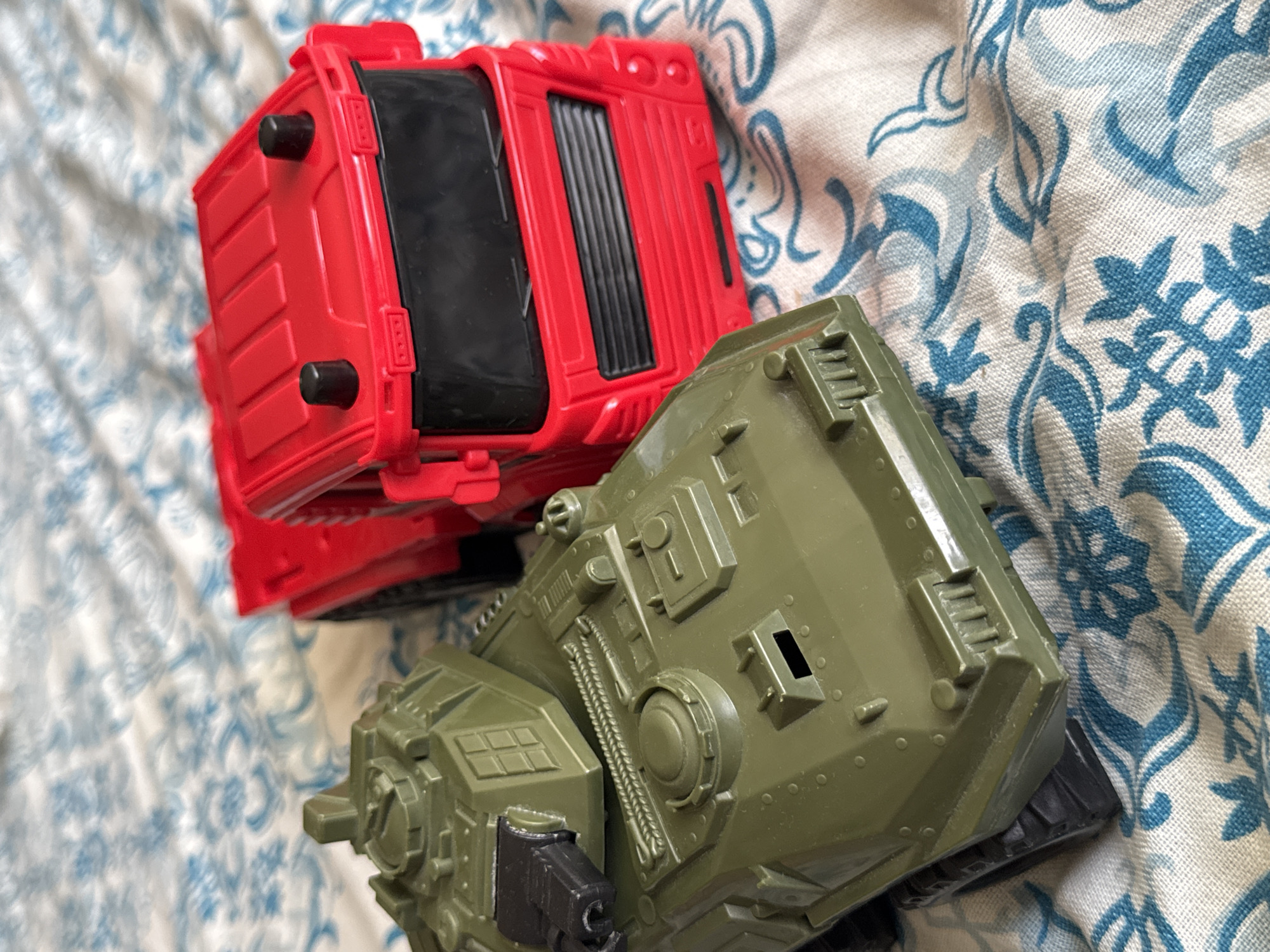 The image shows two toy vehicles, a green tank and a red truck, resting on a bed with a blue and white patterned sheet. The tank is in the foreground on the left, angled towards the viewer. It has a rotating turret with a barrel and a detailed body. The red truck is positioned to the right and slightly behind the tank, facing forward. Its cab is prominent, with a black grille and windshield. The sheet underneath has an abstract floral or swirling pattern in blue and white. The lighting suggests an indoor setting, possibly daytime. The scene is static, with no people or actions depicted.