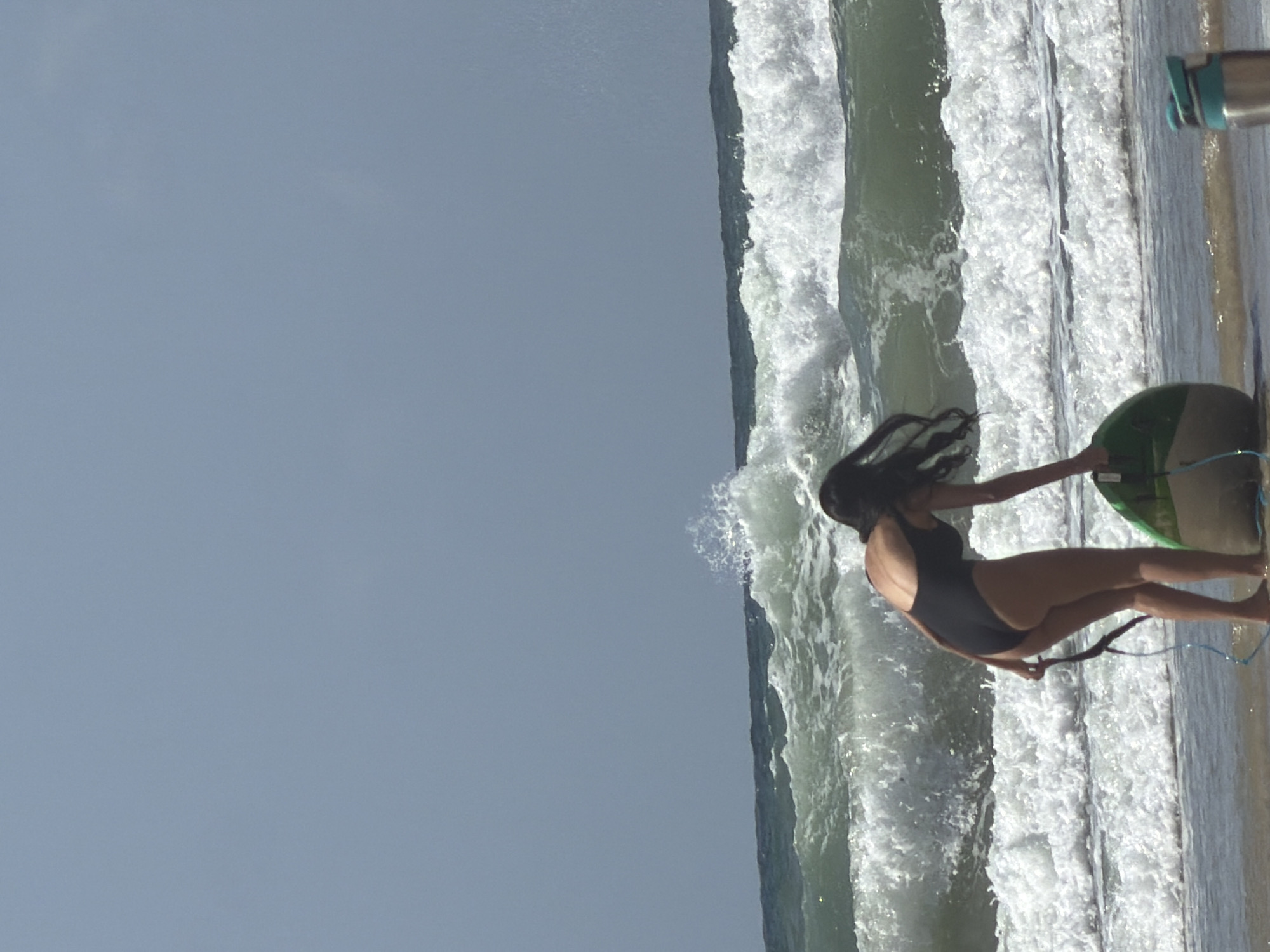 A person in a black swimsuit stands in the surf, holding a green and white surfboard in Tel Aviv, Israel. The sky is blue. The surf is rough.