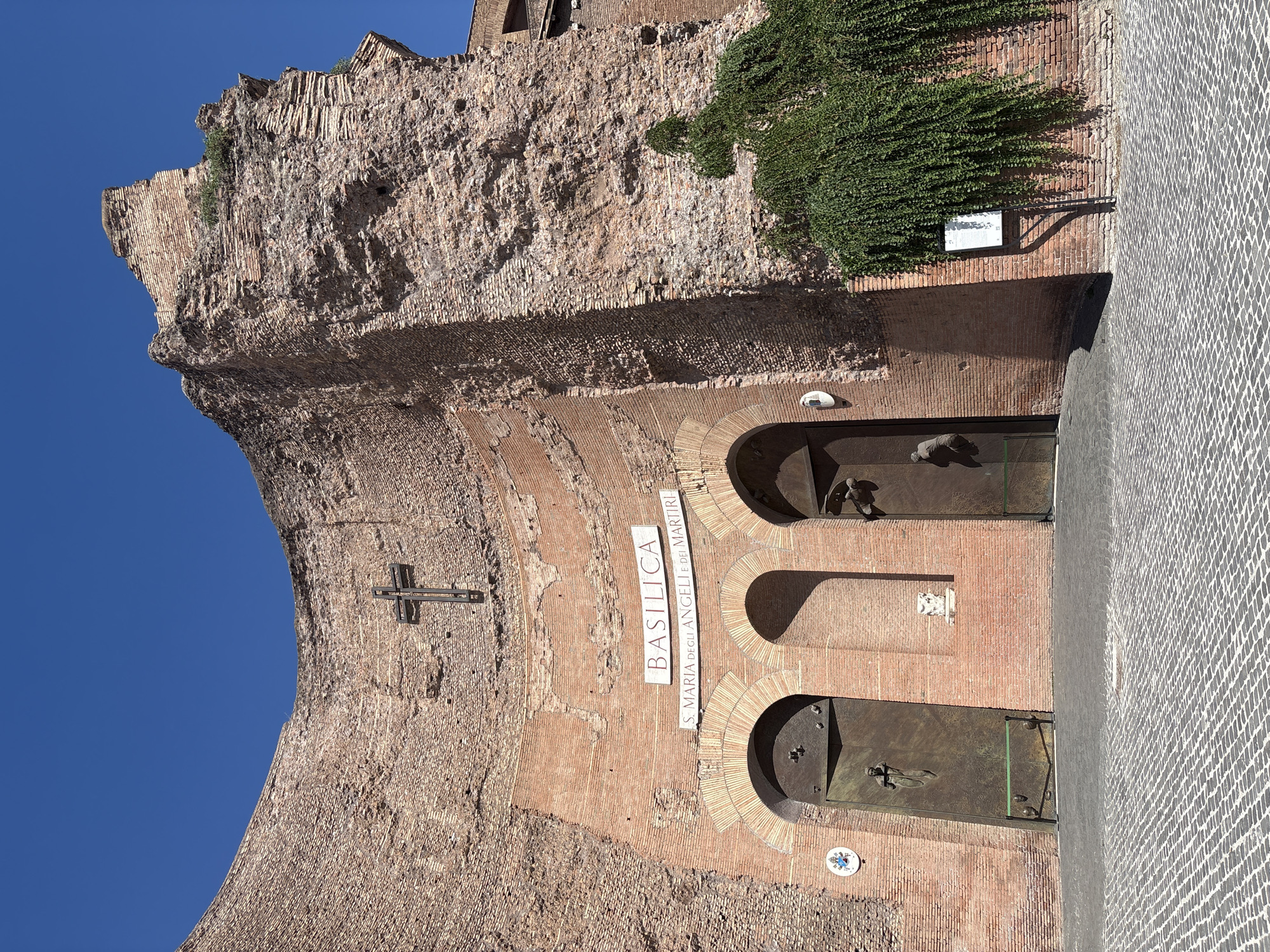 The image shows the facade of the Basilica di Santa Maria degli Angeli e dei Martiri, located in Rome, Italy. The basilica is built within the ruins of the Baths of Diocletian. The facade is made of weathered red brick, with a large, partially ruined arched section at the top. A dark metal cross is mounted on the brickwork above the entrance. Two large, arched bronze doors are visible, with one featuring a relief sculpture of a human figure. Above the doors, signs read "BASILICA" and "S. MARIA DEGLI ANGELI E DEI MARTIRI". Green ivy drapes down the brickwork on the right side of the facade. The ground in front of the basilica is paved with a traditional cobblestone pattern. The sky is a clear, bright blue. There are no people visible in the image.
