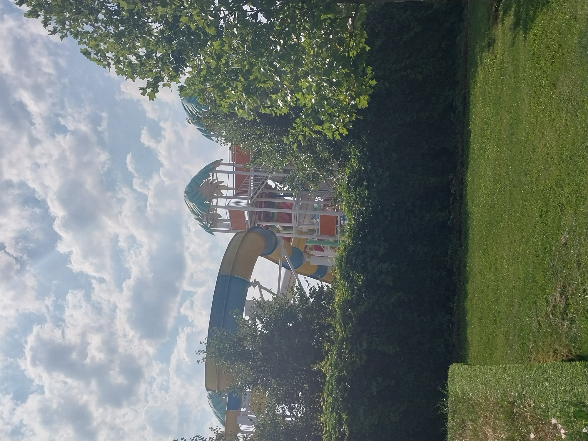 The image depicts a water slide structure at a water park, likely located in Dnipro, Ukraine. The slide is predominantly yellow and blue, with sections of orange visible further back. The structure features a white metal framework supporting the slides and platforms. Several large green trees frame the right side of the image, and dense green foliage forms a hedge in the foreground. The ground in the foreground is covered in green grass. The sky above is a mix of blue with scattered white clouds. There are no people visible in the image.