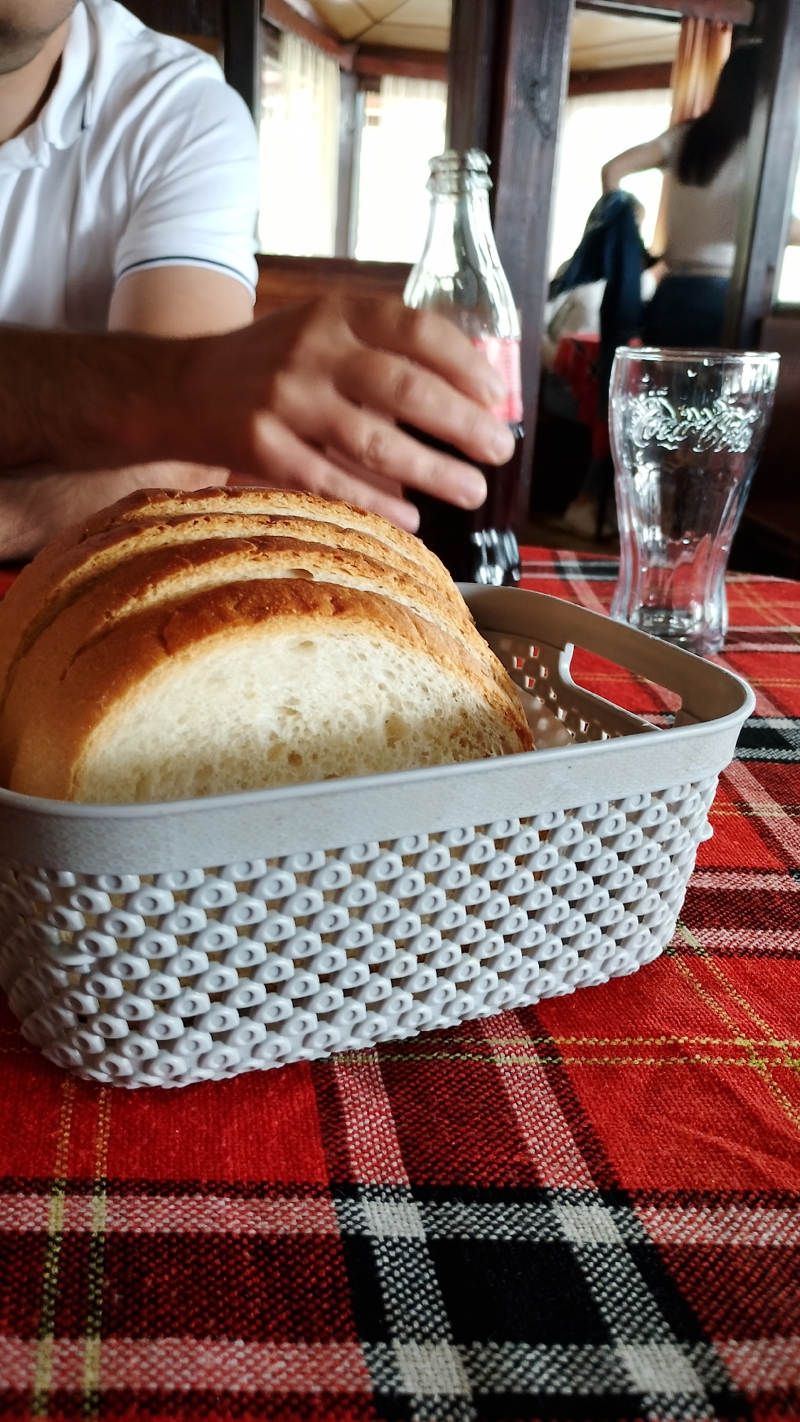 A person's hand is reaching for a glass bottle of Coca-Cola on a table with a red and black plaid tablecloth. In the foreground, a basket of sliced white bread is visible. A clear glass with the Coca-Cola logo is to the right of the bottle. In the background, another person is visible, and the setting appears to be a restaurant or cafe. The location is Pevtsite, Bulgaria.