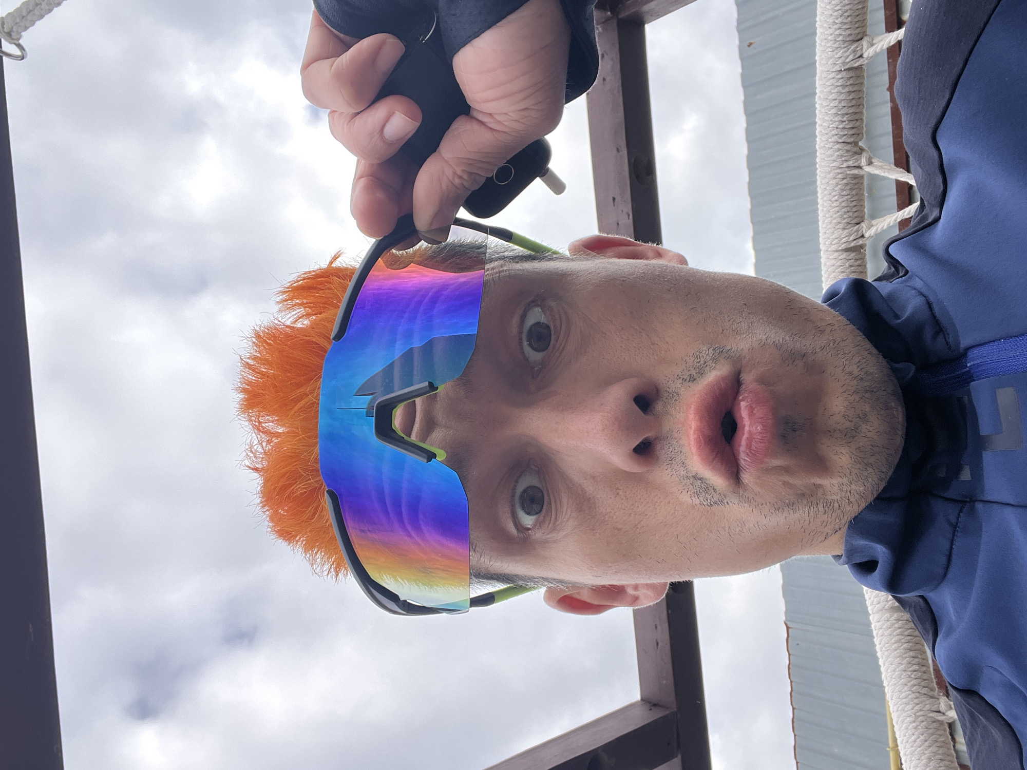 The image shows a close-up, low-angle selfie of a man with bright orange hair and large, reflective cycling glasses. He is wearing a blue and black jacket. His eyes are wide, and his mouth is in a puckered expression. A hand is visible on the right side of the frame, adjusting the glasses. The background appears to be an outdoor setting with a cloudy sky and some wooden structures. There are no specific landmarks visible that would indicate the location is Chaikovsky, Russia.