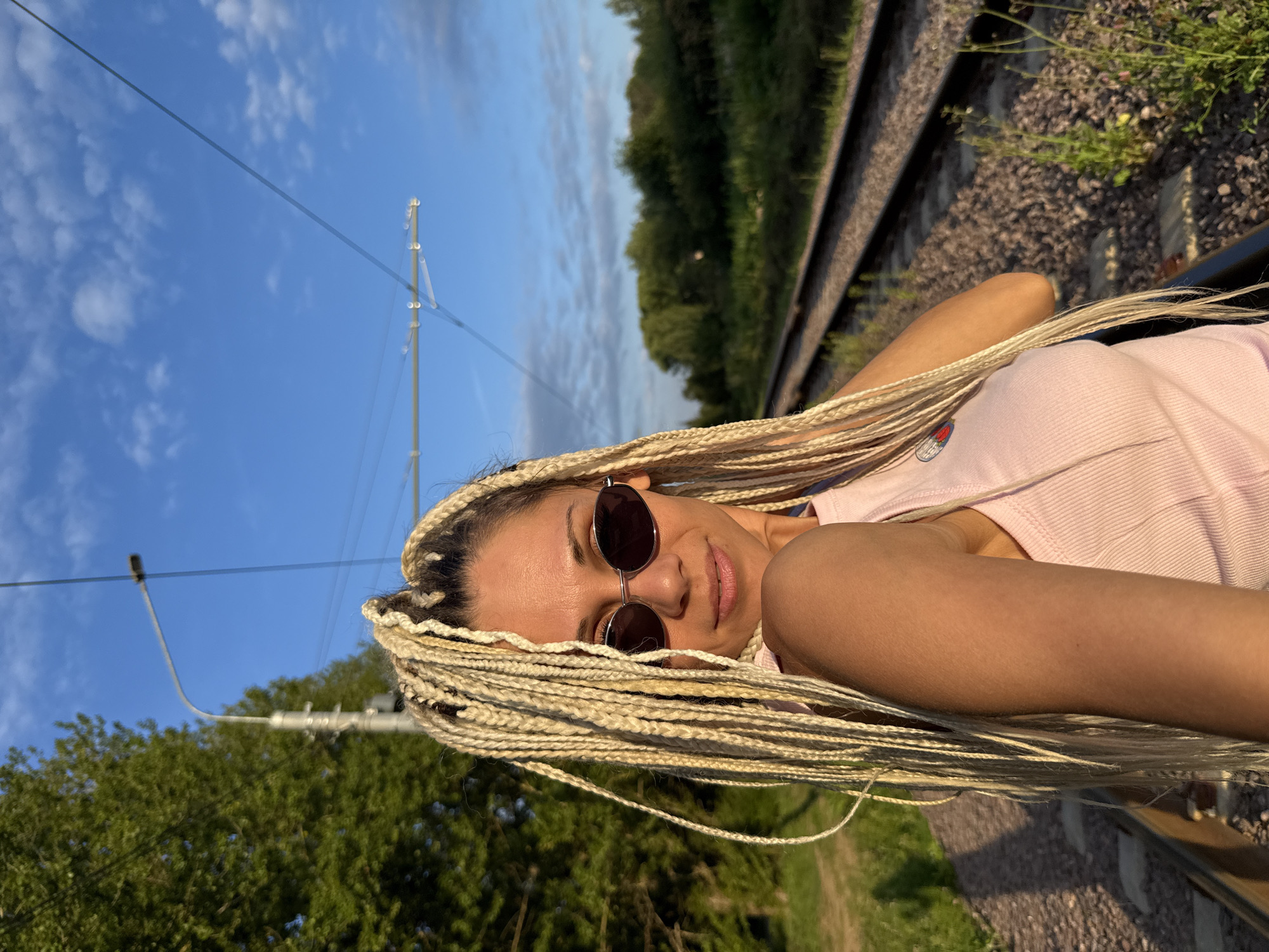 A medium shot shows a woman with blonde braids and sunglasses standing next to a train track. She is wearing a pink tank top and is looking at the camera. The train track runs diagonally through the frame, with trees and vegetation on either side. The sky is blue with a few clouds. The image appears to be taken in Rusanovo, Russia, given the presence of train tracks and typical vegetation for the region.