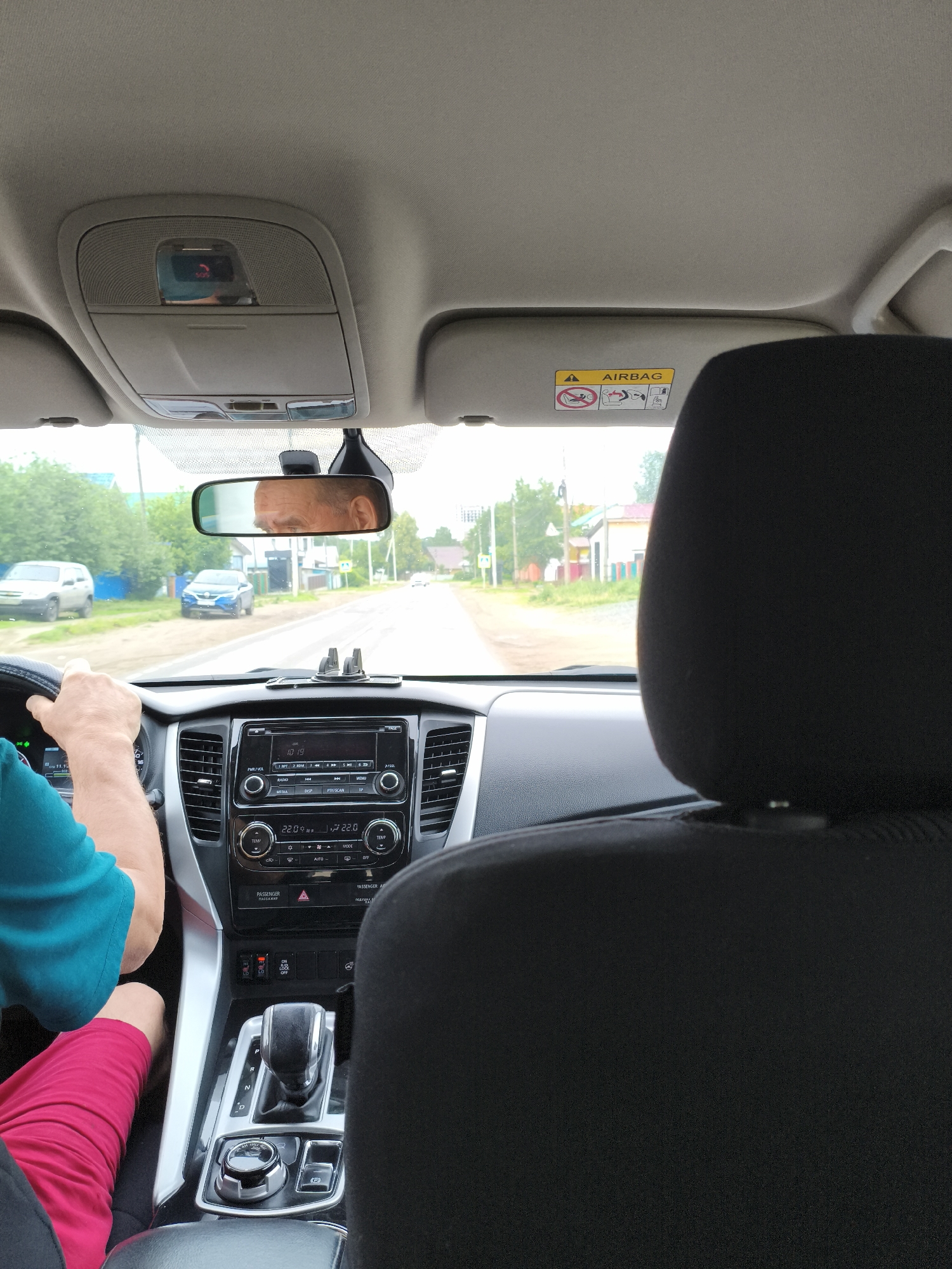 This image, captured from the rear passenger seat of a vehicle, details the car's interior facing forward through the windshield, revealing a rural or suburban street scene.
**Interior:**
The front passenger headrest, upholstered in black fabric, occupies the immediate foreground on the right. To the left, the driver's torso is partially visible, clad in a blue-green short-sleeved shirt and red shorts. The driver's hands, with exposed skin, grasp the steering wheel. The driver's face, an older male with short hair, is reflected in the rearview mirror positioned centrally above the dashboard.
The dashboard features a multi-function steering wheel. The center console includes an infotainment system with a digital display showing "10:19" as the time and "22.0°C" for temperature. Below this are climate control dials and buttons. Further down, an automatic gear selector is set to "P" (Park). To its left, a rotary knob for drive mode selection and an electronic parking brake button are present. Above the gear selector, two red illuminated buttons indicate that both driver and passenger heated seats are active. A black phone mount is affixed to the top of the dashboard.
Above the windshield, the light grey headliner houses an overhead console. This console contains a red illuminated "SOS" button, alongside other controls and a sunglass compartment. The passenger sun visor, also light grey, is folded up and displays an "AIRBAG" warning label with a depiction of a child in a car seat with a cross through it.
**Exterior (viewed through the windshield):**
The vehicle is on what appears to be an unpaved or dirt road that extends into the distance under an overcast sky. The road is flanked by green trees and bushes. Scattered single-story houses with various colored facades (blue, white, yellow/orange) line both sides of the street. Utility poles are visible along the road. A white sedan is parked on the left side of the road closer to the foreground, and a darker blue SUV is parked further down on the same side. A "Pedestrian Crossing" sign is visible on the left side of the road.