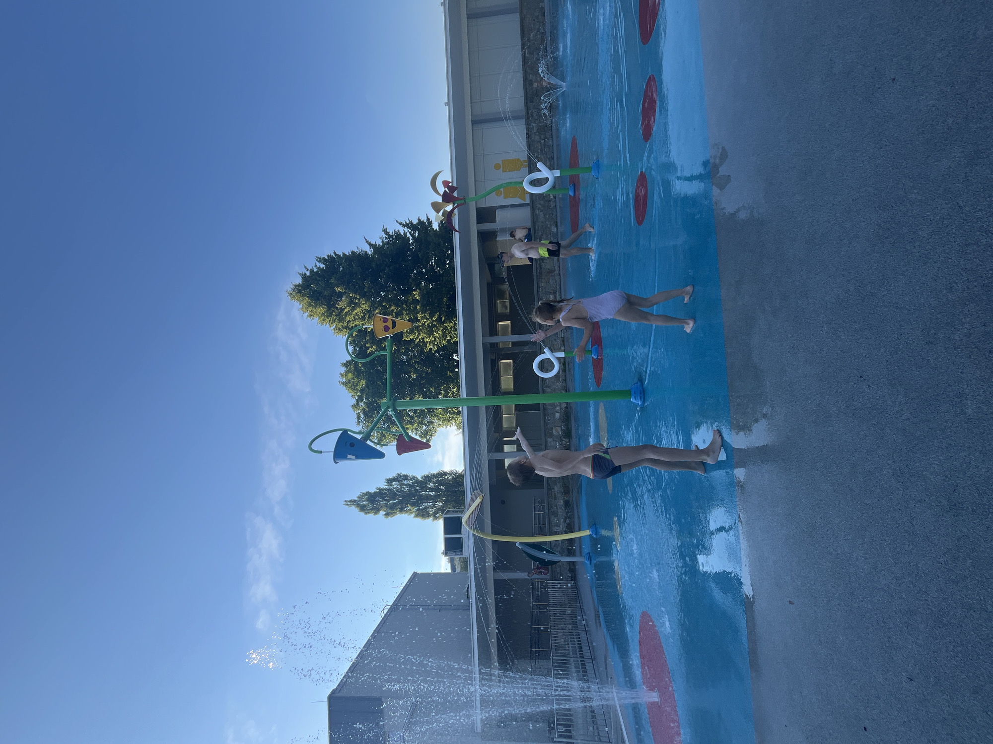 The image depicts a bright, sunny day at a splash pad. In the foreground, a young boy stands in the shallow water, facing away from the camera. He is wearing dark swimming trunks and has his left arm extended. To his right, a young girl in a light-colored swimsuit faces the boy, with her arms raised as if playing. Behind them, a man in yellow swim trunks walks away from the camera.
The splash pad features a bright blue surface with red circular spray pads. Several water features are visible, including a tall green structure with hanging buckets that are either full of water or tipped over, a yellow arc with a spray nozzle, and smaller spray nozzles on white spiral-shaped structures. A large fountain is active on the left side of the image, spraying water high into the air.
In the background, a modern building with a light-colored facade and a covered walkway runs along the edge of the splash pad. Trees with green foliage are visible behind the building. The sky is a clear, vibrant blue. The overall scene is one of children and adults enjoying a recreational water area on a warm day.