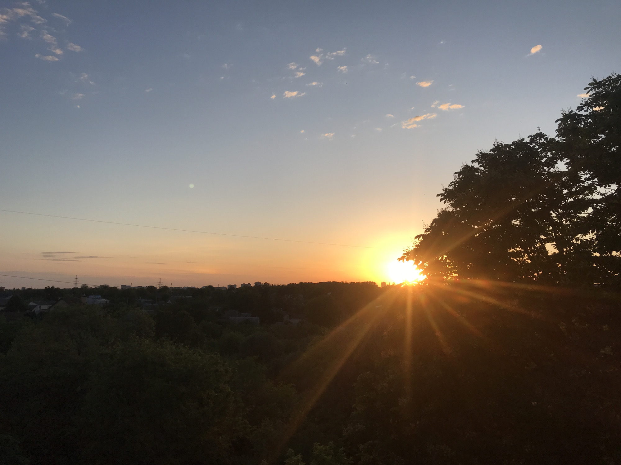 The image depicts a sunset over the city of Kharkiv, Ukraine. The sun is positioned on the horizon, partially obscured by the leaves of a tree on the right side of the frame. The sun's rays are visible, shining through the tree's branches. In the distance, a line of buildings can be seen above the tree line. The sky is a gradient of colors, from blue at the top to orange and yellow near the horizon. Small, scattered clouds are visible in the blue portion of the sky. The foreground is dominated by dark, shadowy trees. A power line is horizontally stretched across the image.