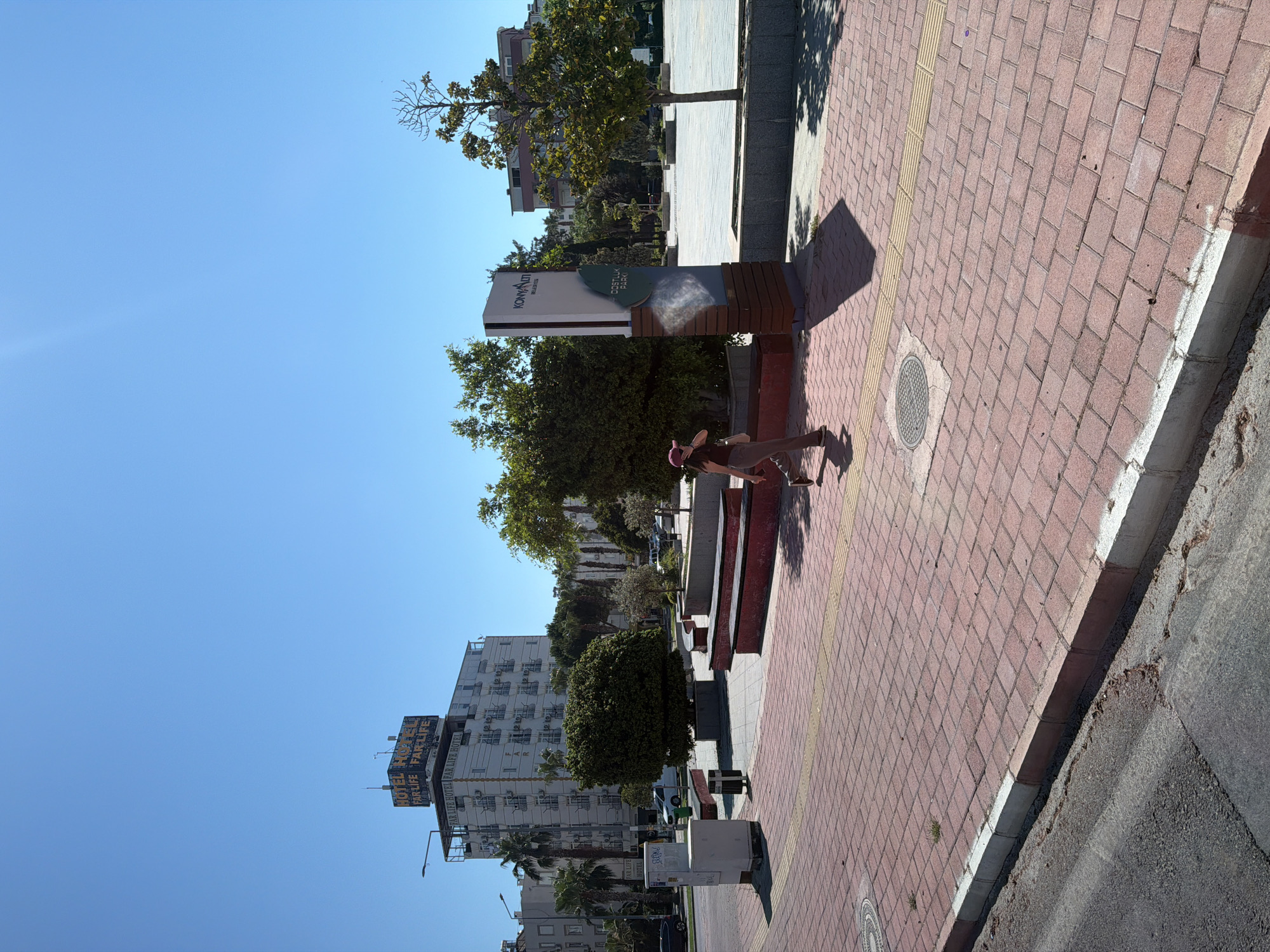 A woman in a pink hat and bikini is captured in a playful pose on a sun-drenched sidewalk paved with red bricks. She is leaning against a modern park bench, her body twisted as if mid-dance or stretch. The scene unfolds outdoors, likely in a public space, suggested by the presence of a signpost with the text "KONYAALTI BELEDIYESI" and a logo, indicating the location is Konyaaltı, Turkey. The signpost is partially obscured by trees, one of which is a small, bare-branched deciduous tree casting a long shadow on the pavement.
Behind the woman and the bench, more trees and a glimpse of a multi-story building with white balconies are visible. Further in the background, another tall, white building prominently displays a large sign reading "FAR LIFE HOTEL," with the words "PARK LIFE HOTEL" also visible, suggesting a hotel or resort area. The clear blue sky and strong shadows indicate it is a sunny day, likely late morning or early afternoon. The woman’s posture and the bright environment evoke a sense of leisure and enjoyment. The sidewalk also features a yellow tactile paving strip and a manhole cover. The overall impression is of a warm, bright day in an urban environment, possibly a tourist area.