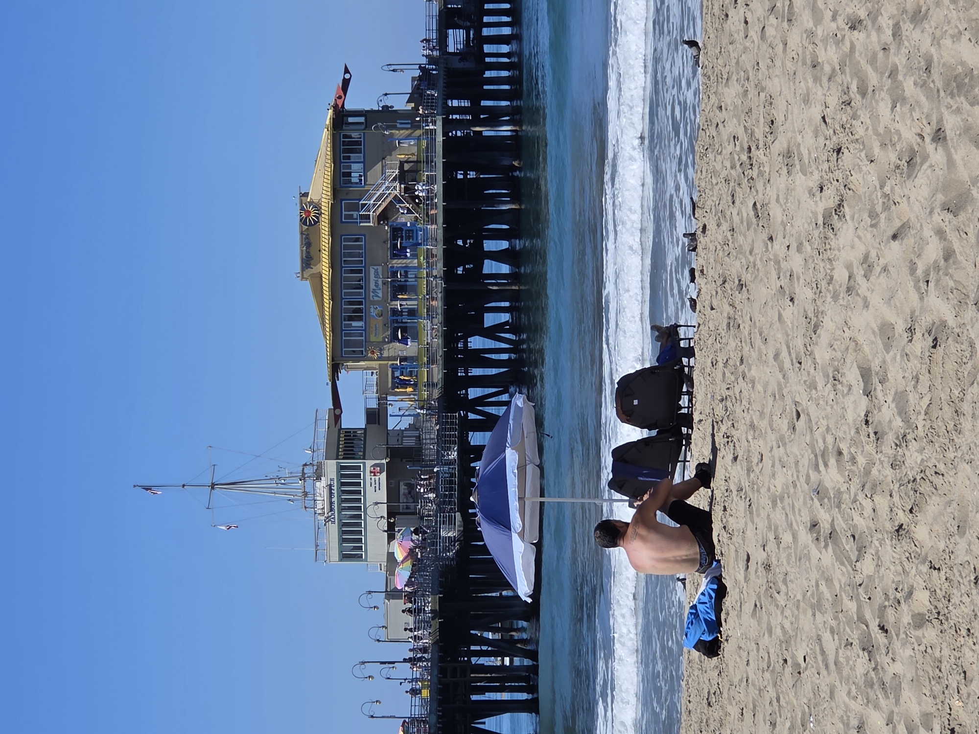 The image shows a view of the Santa Monica Pier in Santa Monica, California, United States of America. In the foreground, a man is sitting on the sand facing away from the camera, with a blue and white beach umbrella and two chairs next to him. There is a blue bag on the sand to his left. The ocean is visible behind him with waves gently rolling onto the shore. The pier extends into the ocean, featuring various buildings and structures, including a building with a yellow roof and a "Harbor Office" sign. Numerous people are visible on the pier, and lampposts line its edge. The sky is clear and blue.
