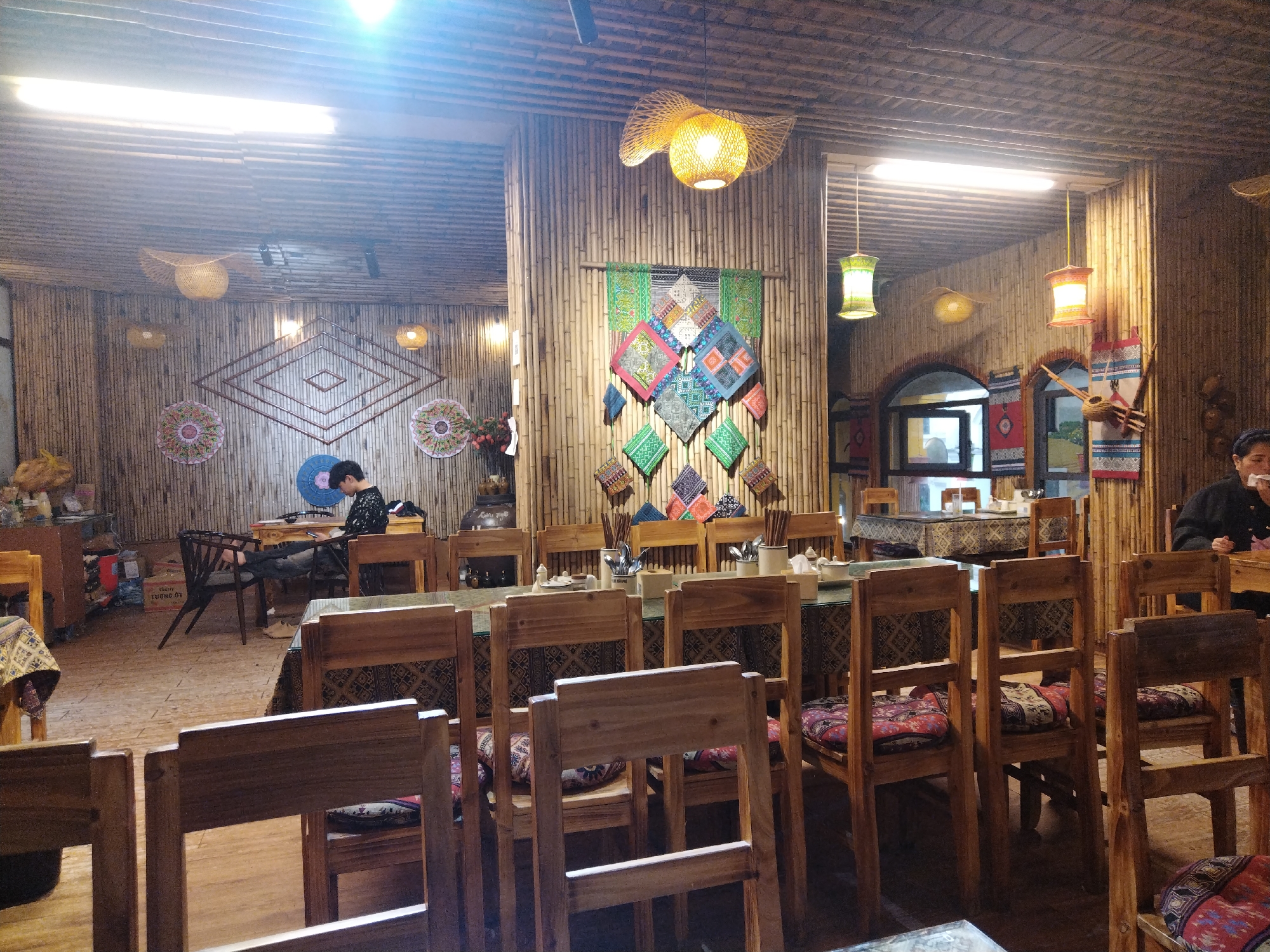 The interior of a restaurant in Sa Pa Ward, Vietnam, features tables set with tableware and decorated with tablecloths, surrounded by wooden chairs with patterned cushions. Walls and columns are clad in bamboo. Hanging decorations include woven lamps and colorful textile art. A person is seated at a table in the background on the left, and another person sits at a table to the right. Chopsticks and serving utensils are placed on the tables.