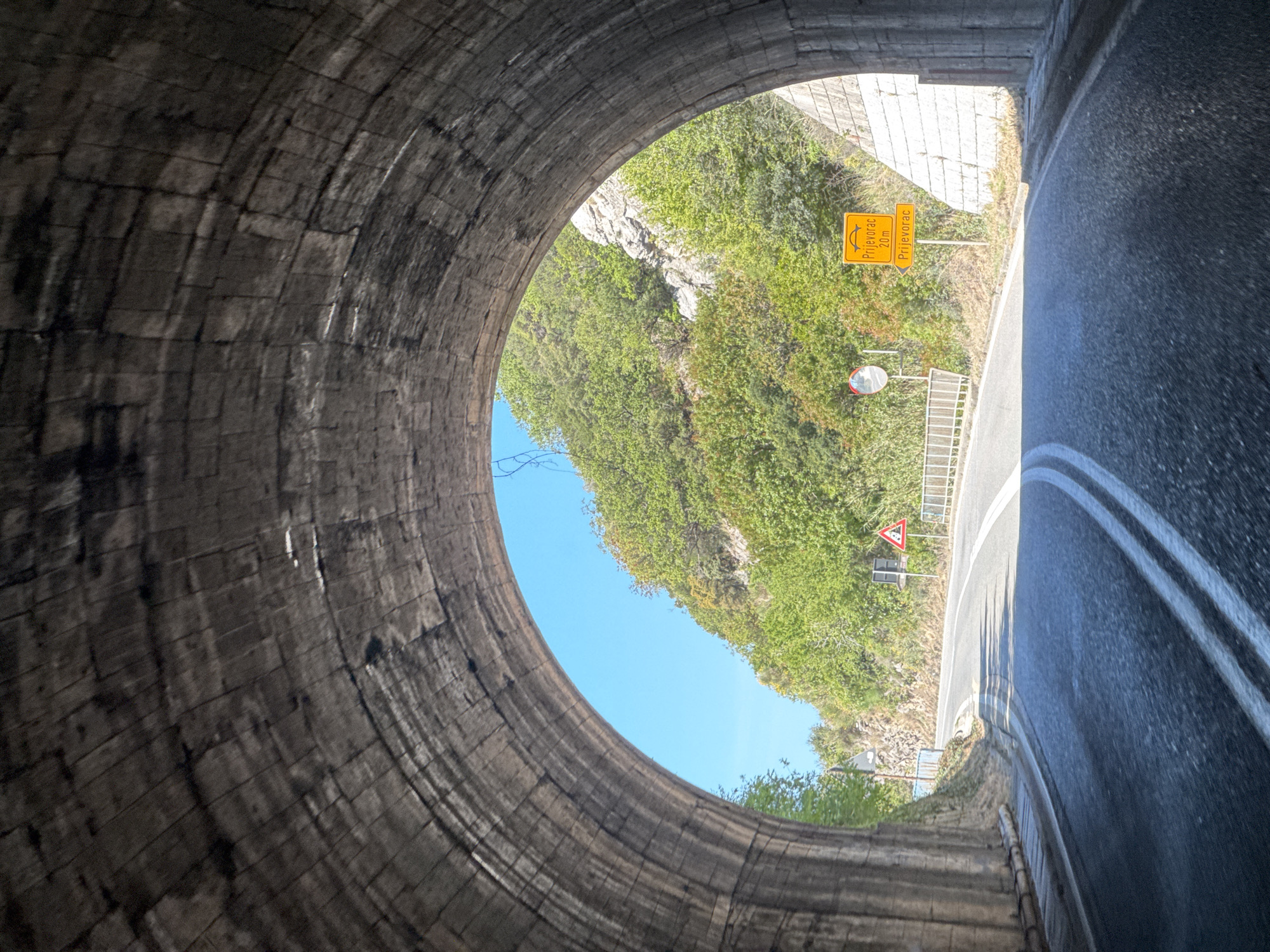 The image shows the view from inside a stone tunnel looking out onto a road and hillside. The road, marked with white lines, curves to the right. A yellow sign indicating "Prijevorac 20m" is visible on the left side of the road, along with other road signs, including one shaped like a triangle. A lush, green hillside covered in vegetation rises behind the road, visible through the tunnel opening. The sky above the hillside is blue. The tunnel walls are made of dark, aged stone blocks. The image was taken in the city of Androvići, Montenegro.