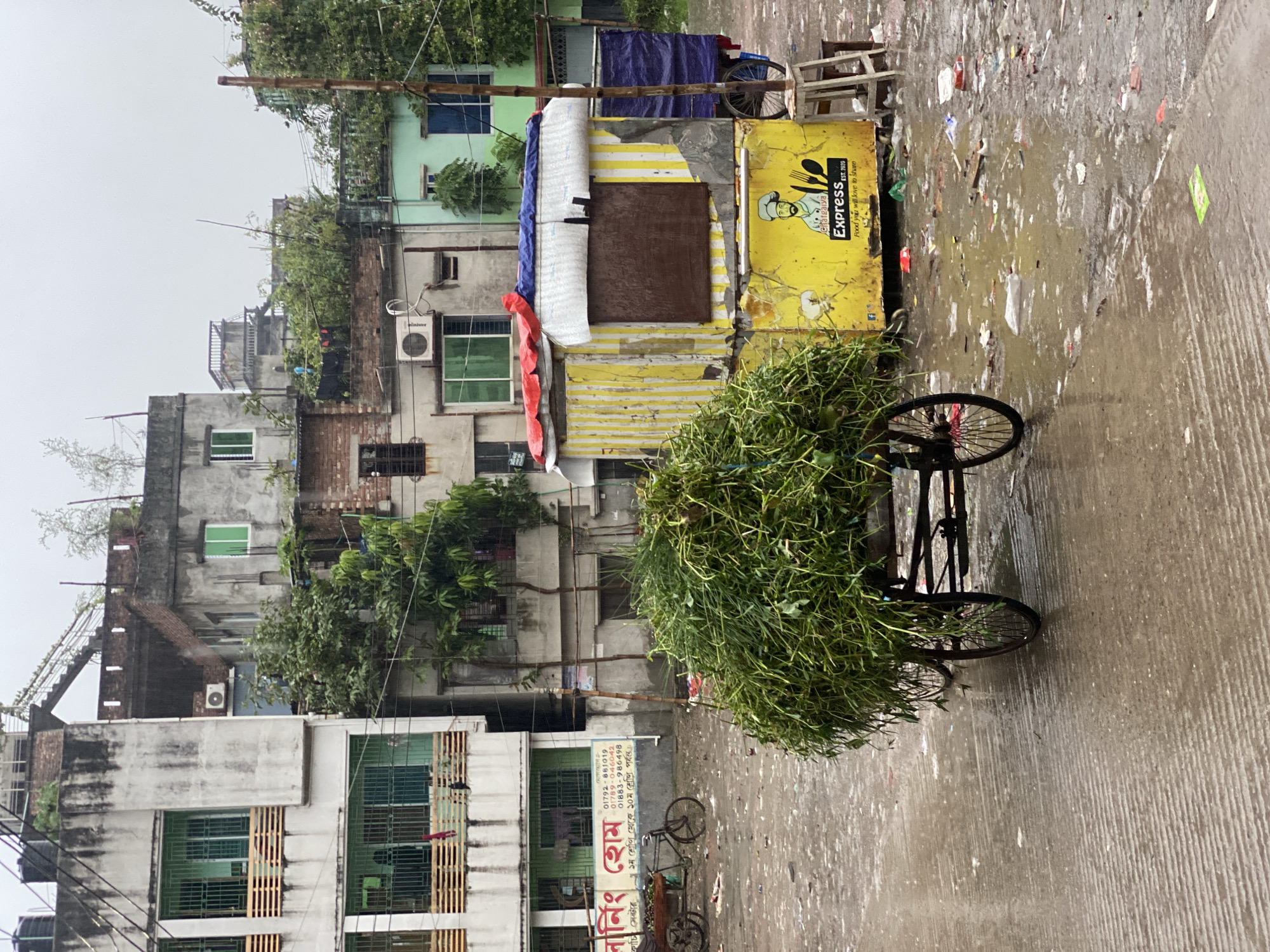 The image depicts a street scene in Dhaka, Bangladesh, likely after rainfall, judging by the wet pavement and general overcast atmosphere. A three-wheeled cart loaded with leafy green vegetation dominates the foreground. Behind the cart is a food stall painted yellow and with the name "Chatgaiya Express". The stall has corrugated metal siding and is partially covered with a blue tarp and white striped material. The background features a collection of densely packed buildings of varying heights and architectural styles, reflecting a typical urban environment in Dhaka. A sign is visible on one of the buildings which says "বানিং হোম" and includes telephone numbers. There's litter and debris scattered on the wet street, indicating a lack of immediate sanitation.