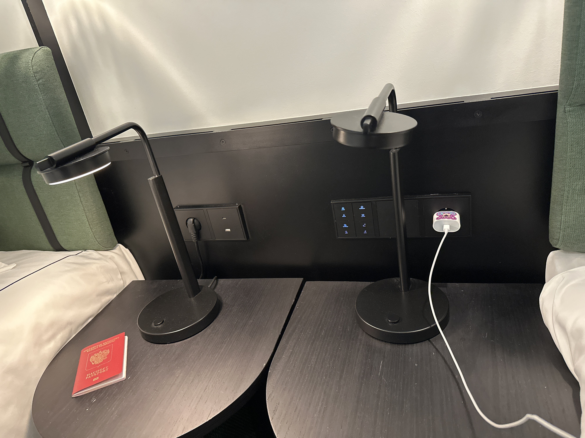 The image depicts a bedside scene in a hotel room in Tbilisi, Georgia. Two black bedside tables are positioned on either side of a bed with white linens and green cushions. Each table has a black desk lamp.
On the left table, there is a red Russian passport. The lamp is switched on.
On the right table, a white charging cable with a decorative cartoon-character plug is plugged into a wall outlet. The lamp is not switched on.
The headboard behind the tables is black and features electrical outlets and light control panels. The wall is white.