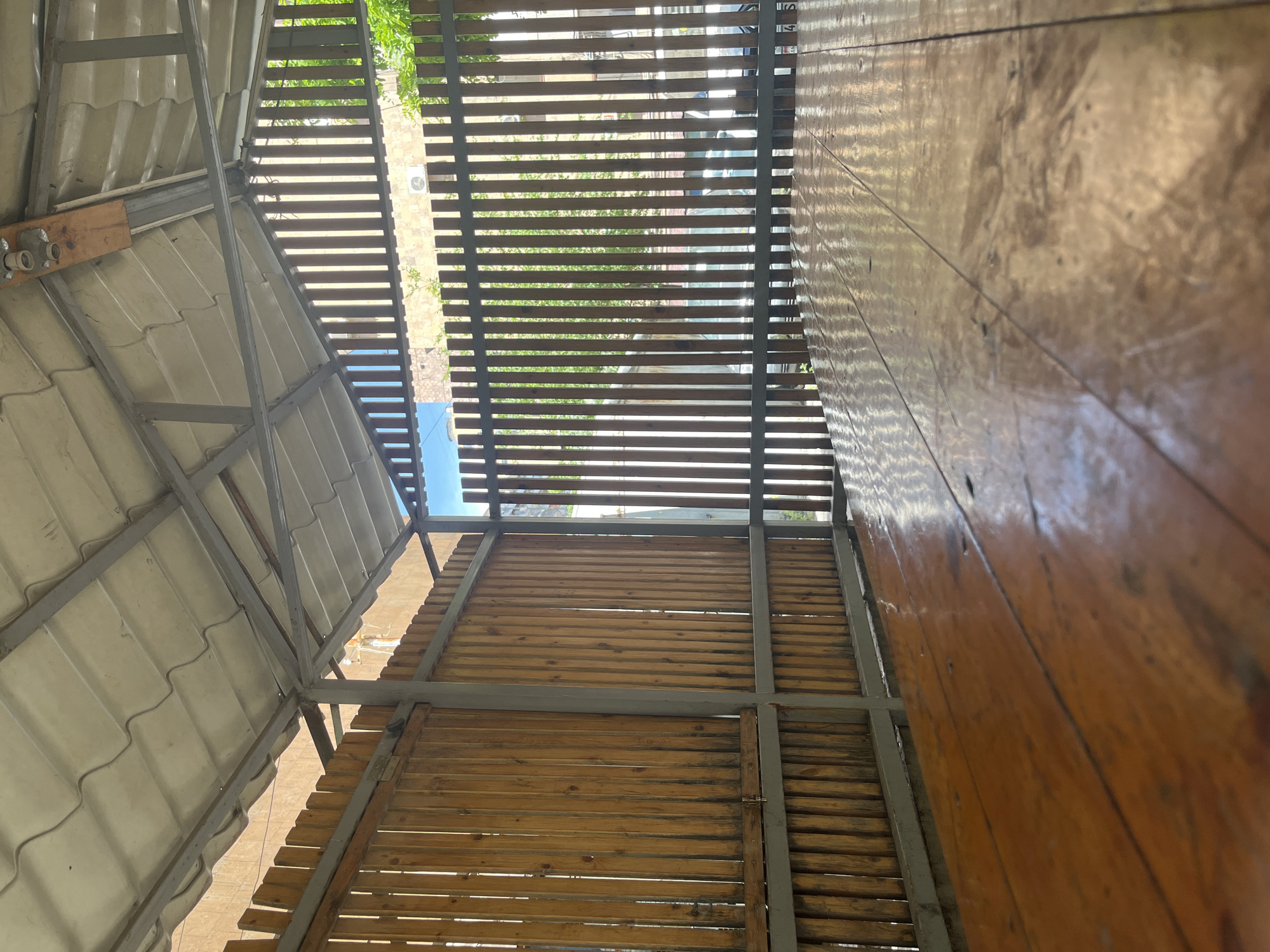 The image shows the interior of a structure, likely a balcony or covered patio, in Yerevan, Armenia. The scene is viewed from below, looking up towards the roof and walls.
Roof: The roof is constructed of corrugated white panels supported by a grey metal frame.
Walls: The walls are constructed of horizontal wooden slats, also supported by a grey metal frame. Some sections have narrower slats, forming a sort of louver or blind. The wooden slats have a glossy, varnished finish.
Environment: Through the gaps in the slats, glimpses of the outside world are visible, including greenery (possibly trees) and buildings. The sky is visible through the gaps in the upper part of the image.
General: The overall impression is of a covered outdoor space that provides shade and privacy. The perspective is unusual, looking upwards, which emphasizes the structural elements of the enclosure. There are no people visible in the image.