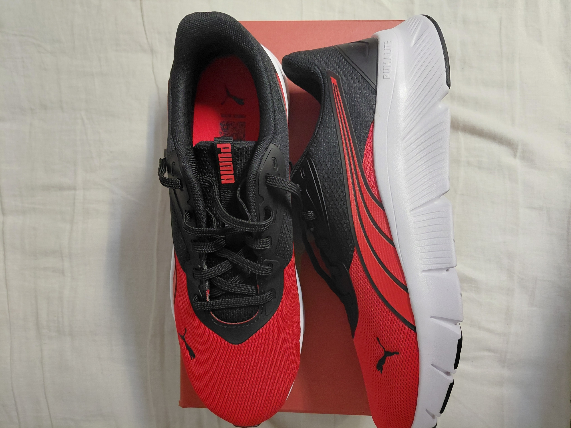 The image shows a pair of Puma running shoes in red and black colors. The shoes are placed on top of a red box, which is set on a white fabric surface. The shoe on the left faces the camera, displaying the laces, red mesh material, and black Puma logo. The right shoe is placed sideways, showing the white sole and black and red side design. "PUMALITE" is printed on the white sole. The image provides a product shot of the shoes. There is no indication of Sofia, Bulgaria in the image.