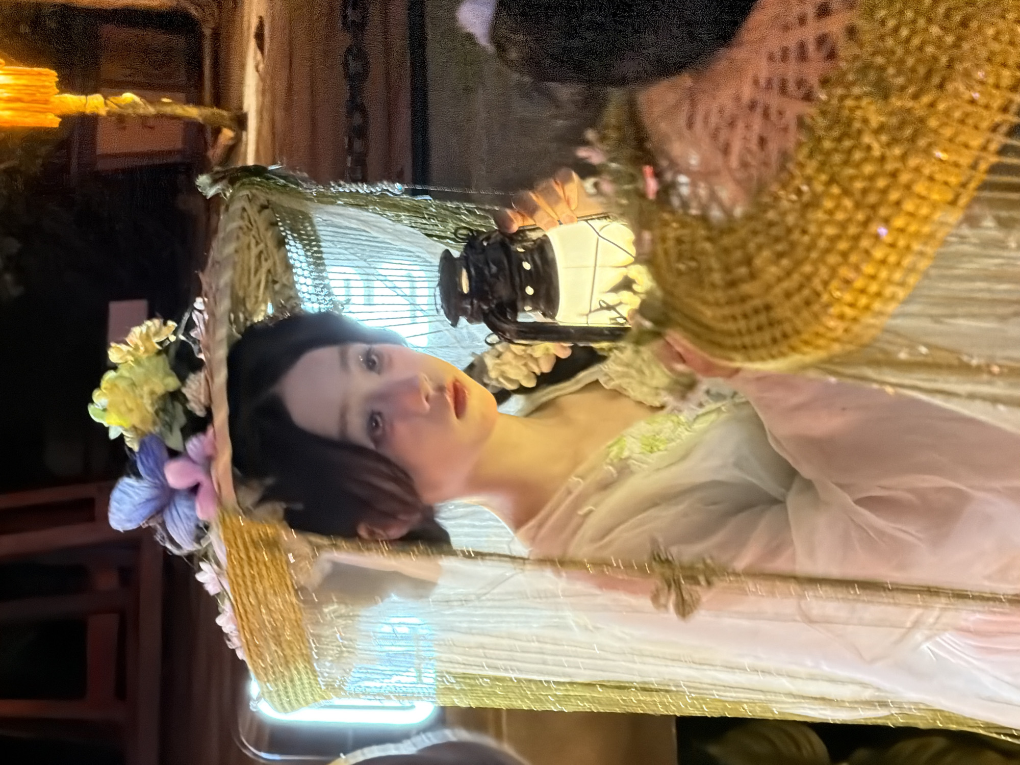 The image shows a young woman in traditional Chinese attire holding a lantern. She is wearing a wide-brimmed straw hat decorated with flowers. Her dress is light-colored and flowing, with delicate embroidery. The woman has dark hair pulled back and a serious expression on her face. She is holding a lit lantern in her right hand, which illuminates her face and the front of her dress. The background is dark and appears to be an outdoor setting at night, possibly a street or courtyard in Yuzhong District, China, given the presence of traditional architecture and lanterns. The overall mood is ethereal and somewhat mysterious.