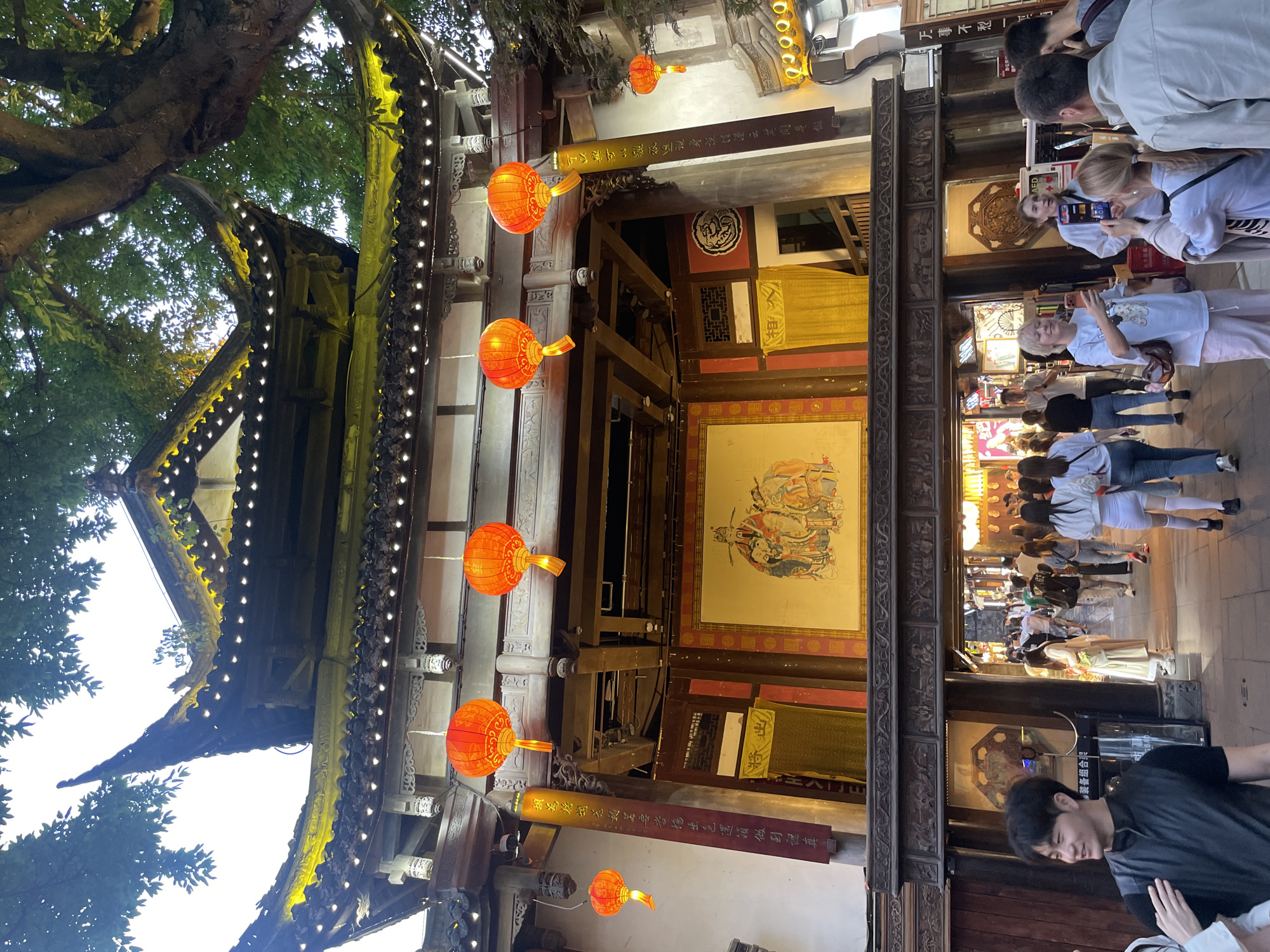 This image shows a view through an ornately decorated building, likely a temple or historical structure in the Wuhou District, China. The architecture features traditional Chinese elements like tiered roofs, intricate wooden carvings, and red lanterns. Strings of lights accent the rooflines. Inside the building, there's artwork on the walls, possibly depicting historical or religious scenes. The foreground shows parts of the building's framework and decorations. Through the building, people are walking around, taking pictures, and generally observing the scene, suggesting a tourist destination or busy public space. One person in the foreground wears a black t-shirt and has their head down. The overall impression is a mix of traditional architecture and a contemporary tourist environment.