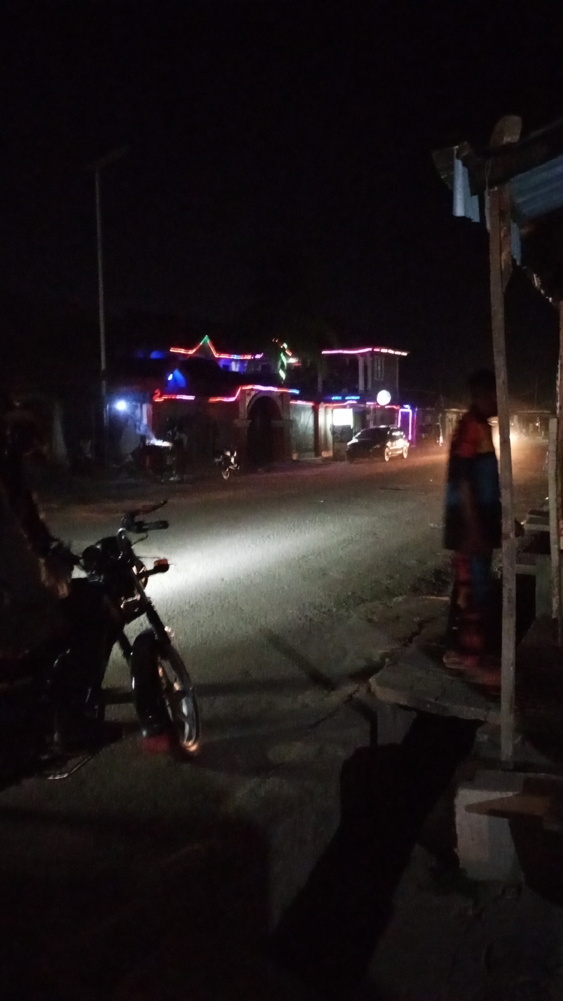 The image shows a night scene in Sabon Tasha, Nigeria. A motorcycle with its headlight on is parked on the left side of a paved road. In the background, a two-story building is decorated with red, blue, and green lights. A car and another motorcycle are parked near the building. A person is standing near a wooden structure on the right side of the road. A streetlight is visible in the background. The environment is dark.