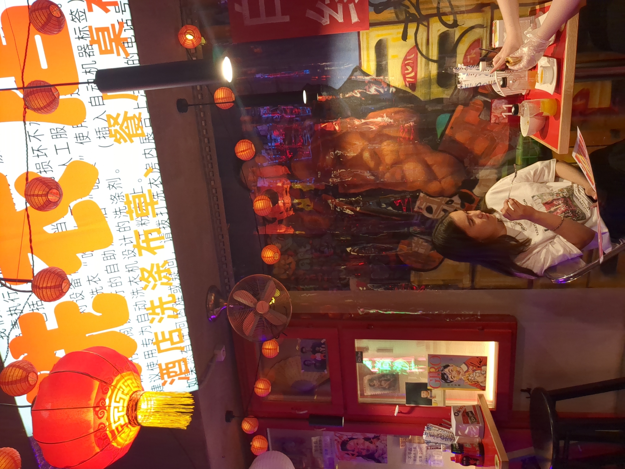 The image displays an interior scene of a themed eatery located in Tyumen, Russia. Two individuals are present within the frame. A young woman, characterized by long, dark hair and a white short-sleeved t-shirt with a graphic print, is seated at a red-topped table on the left, holding a fork towards her mouth. From the right side of the frame, the gloved hands and forearms of a second individual are visible, holding a small food item over the table, indicating an action of serving or food preparation.
The establishment's decor features a prominent East Asian theme. Multiple red Chinese lanterns, including a large, elaborate one with a yellow tassel, are suspended from the ceiling. A large, illuminated sign in the upper background displays Chinese characters, notably "酒店洗" (hotel wash) and "餐" (meal/restaurant), along with smaller script, against a white backdrop.
The walls are adorned with extensive, colorful murals depicting stylized figures and abstract patterns in shades of brown, orange, and yellow. Sections of the walls and a window frame on the left are painted in a solid red. Various framed pictures and posters are displayed, including a magazine cover titled "GIRO" featuring a cartoon figure, visible within a red-framed display case on the left. A wall-mounted oscillating fan is positioned in the upper central area of the room.
The red-topped table where the individuals are situated holds a collection of dining items: a white napkin dispenser, several clear bottles (one containing a yellow liquid), small white bowls, and a paper menu or flyer. A table marker bearing the number "11" is also visible. The ambient lighting is provided by the lanterns, overhead spotlights, and a distant neon glow from a rear area. A dark, round stool is positioned at a smaller counter on the far left.