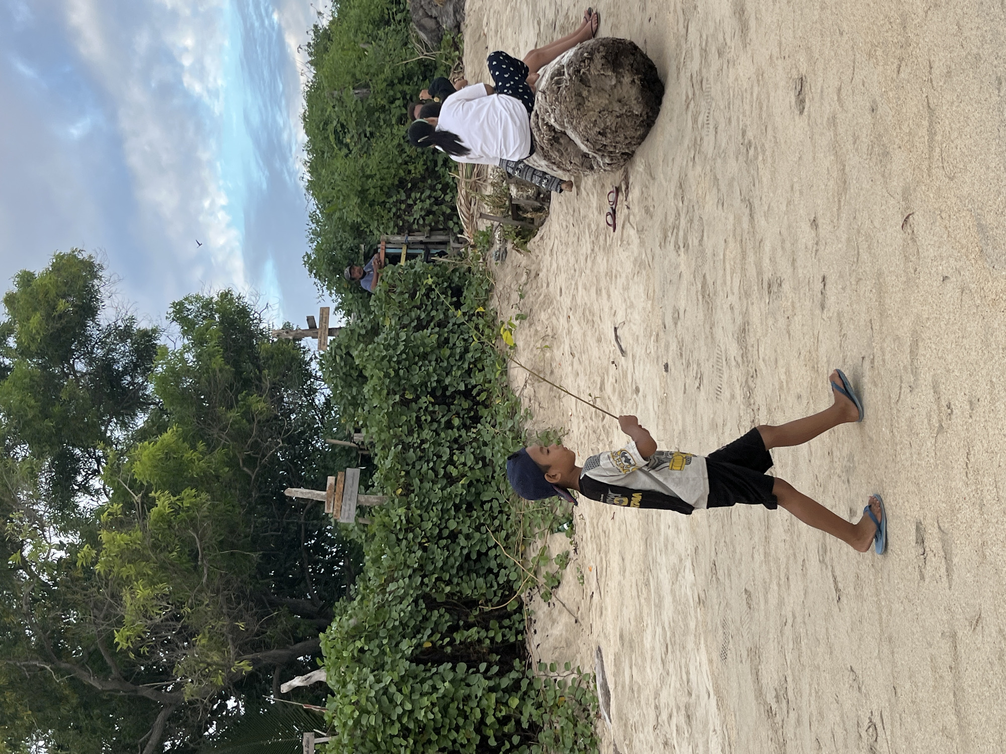A young boy wearing a dark blue baseball cap, a t-shirt with a black and white graphic design, and black shorts walks across a sandy beach. He is holding a thin stick in his right hand. The boy is wearing blue flip-flops. In the background, a woman with dark hair pulled back is sitting on a large, rough log, wearing a white t-shirt and patterned shorts. Another person is seated next to her. Behind them, dense green vegetation covers the landscape, with several wooden crosses visible. Further back, a man is standing near a wooden structure with a fence. The sky above is cloudy with patches of blue. The scene appears to be located on a beach in Kuta, Indonesia, characterized by its sandy terrain, coastal vegetation, and the presence of cultural markers like the crosses.